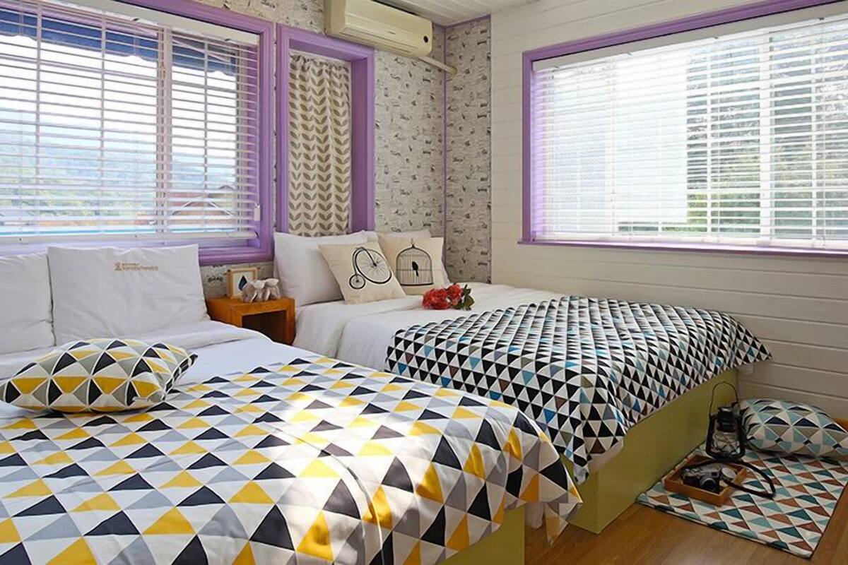 Quarto com duas camas, roupa de cama estampada, mesa de cabeceira e janela com persianas.