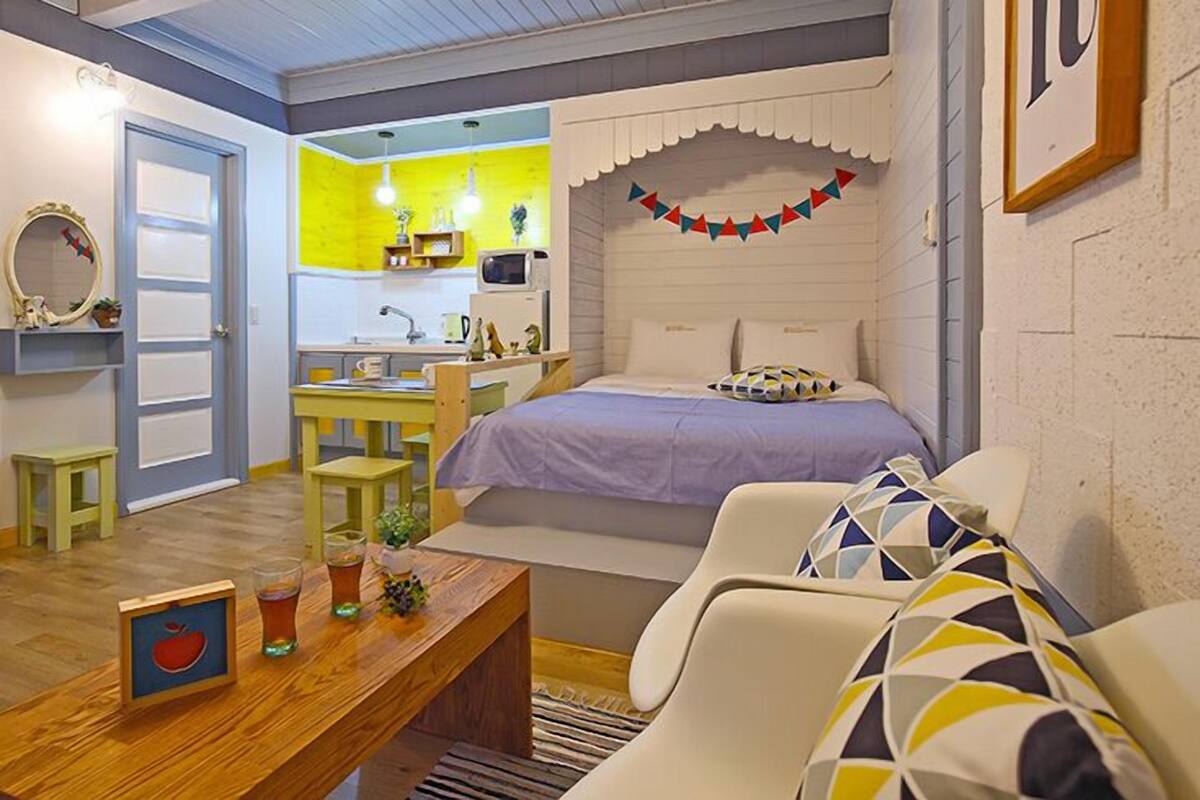 Quarto compacto com cama, uma mesinha com bebidas, uma cadeira e uma área de cozinha compacta.