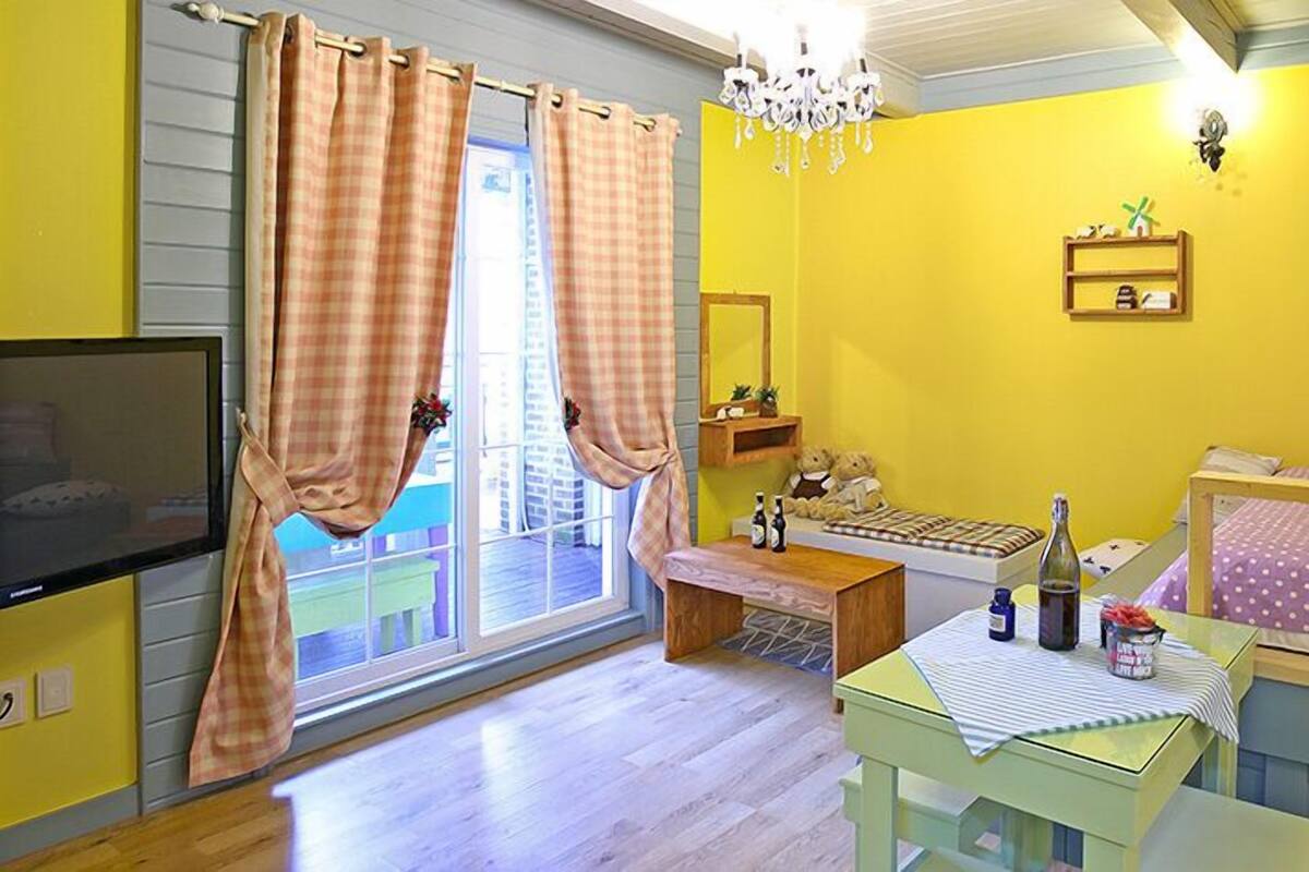 Quarto com parede amarela, cama de madeira, mesa pequena com garrafas, um ursinho de pelúcia e uma televisão.