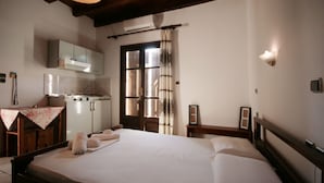 A bedroom with a large bed, a wooden headboard, a nightstand, a small table with a floral tablecloth, and a kitchen area with cabinets and a stove. - Chania Rooms (Chania)