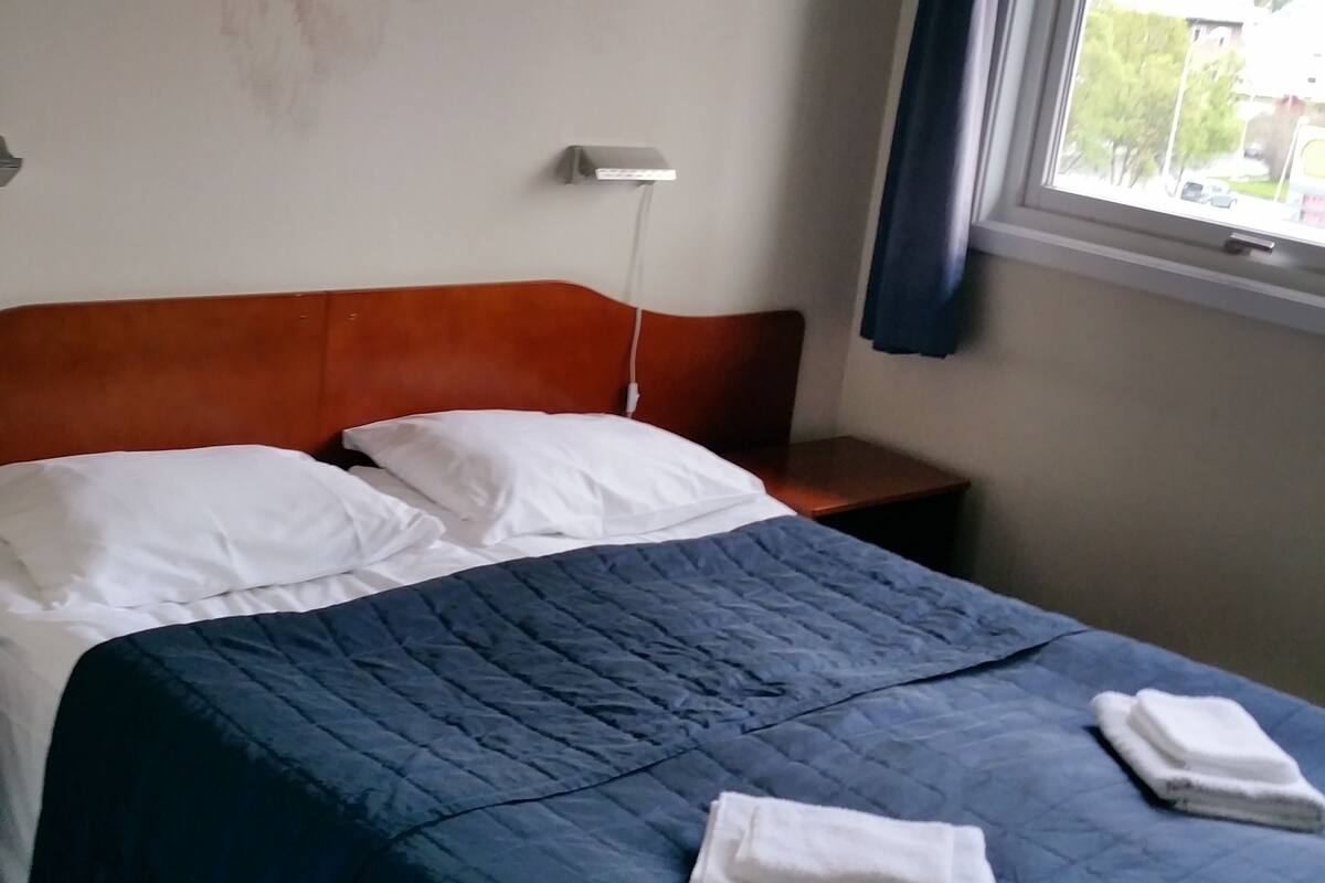 A hotel room with a bed, a nightstand, a wall mural, and a window with curtains.
