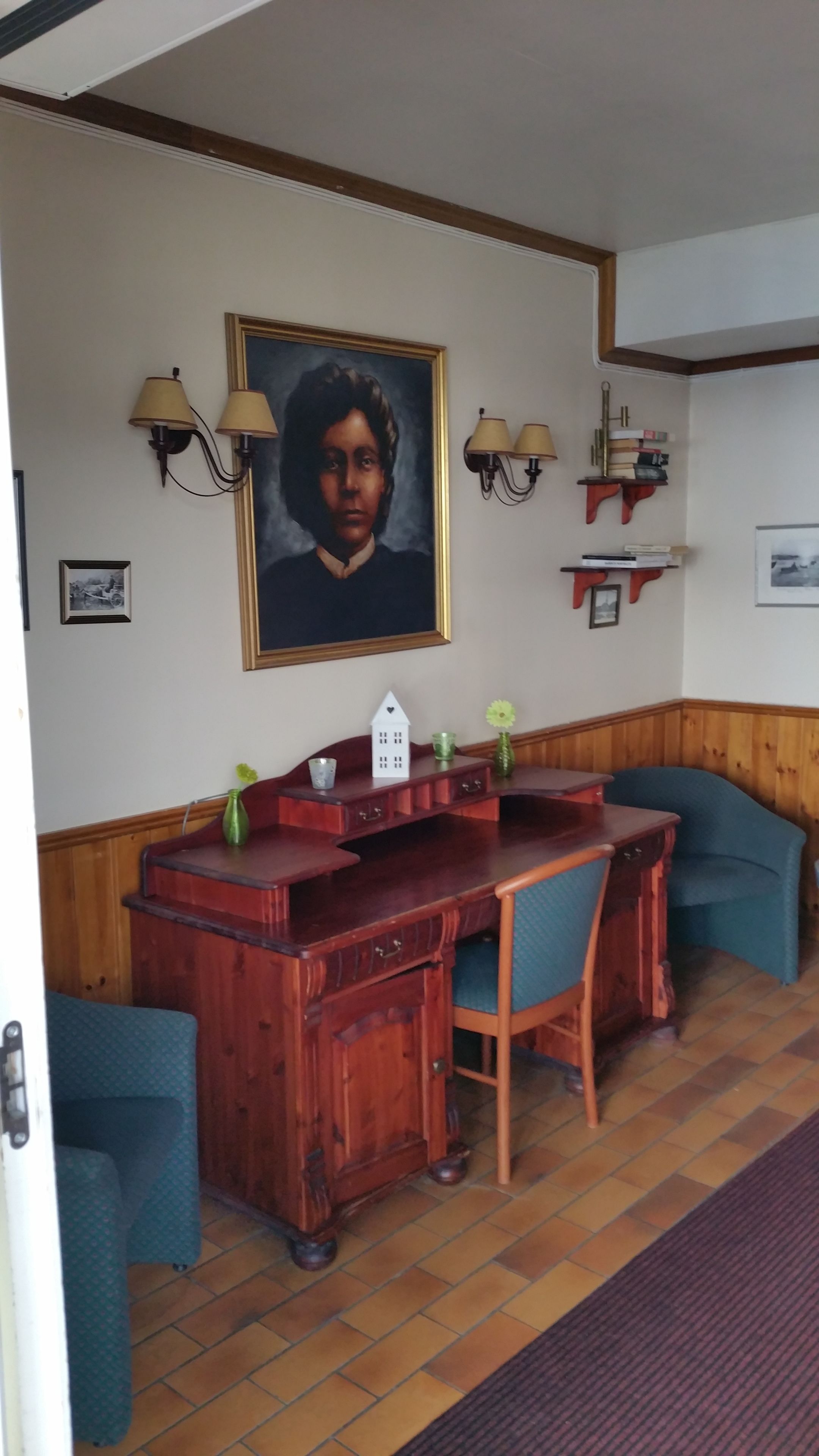 Habitación con escritorio de madera, dos sillas, un retrato enmarcado, lámparas fijadas a la pared y una estantería con libros y un jarrón.