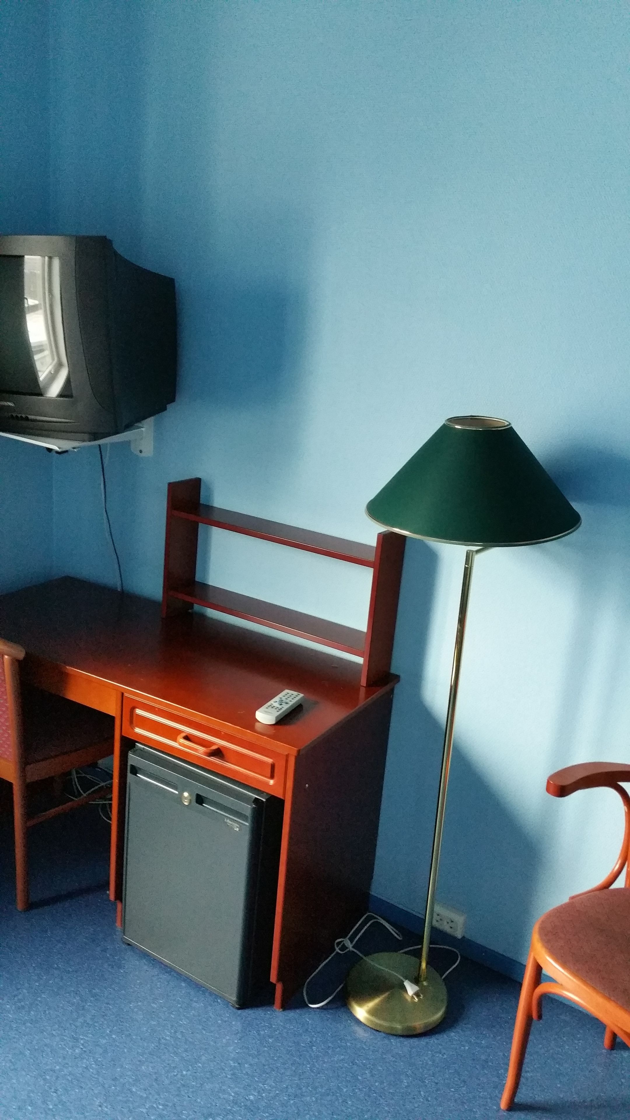 A room with a wooden desk, a small black refrigerator, a green floor lamp, a television mounted on the wall, and a red chair.