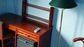 Una habitación con un escritorio de madera, un refrigerador negro pequeño, una lámpara de pie verde, un televisor montado en la pared y una silla roja.