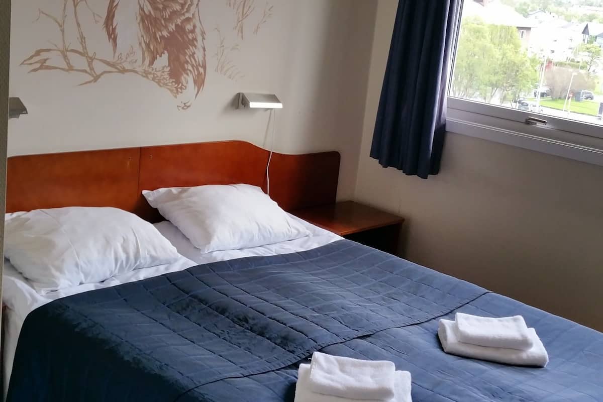 A hotel room with a bed, pillows, a headboard, a wall mural, a window with curtains, and a view of the outside.
