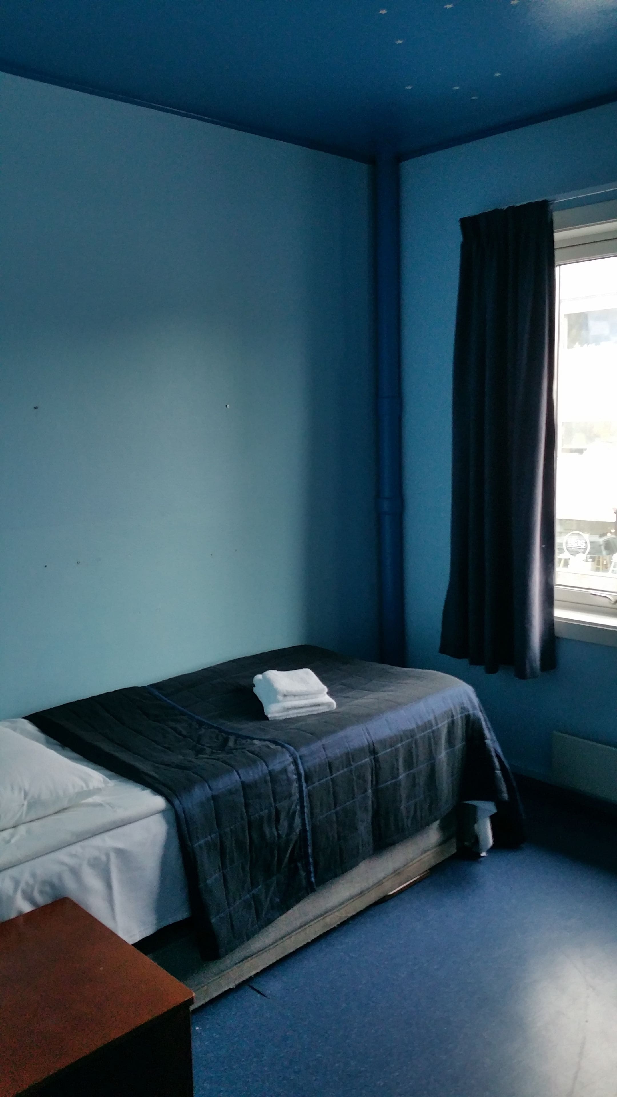 A small, single-bed room with a dark blue wall, a window with curtains, and a visible bed with a white pillow and a folded blanket.