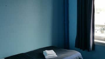 A small, single-bed room with a dark blue wall, a window with curtains, and a visible bed with a white pillow and a folded blanket.