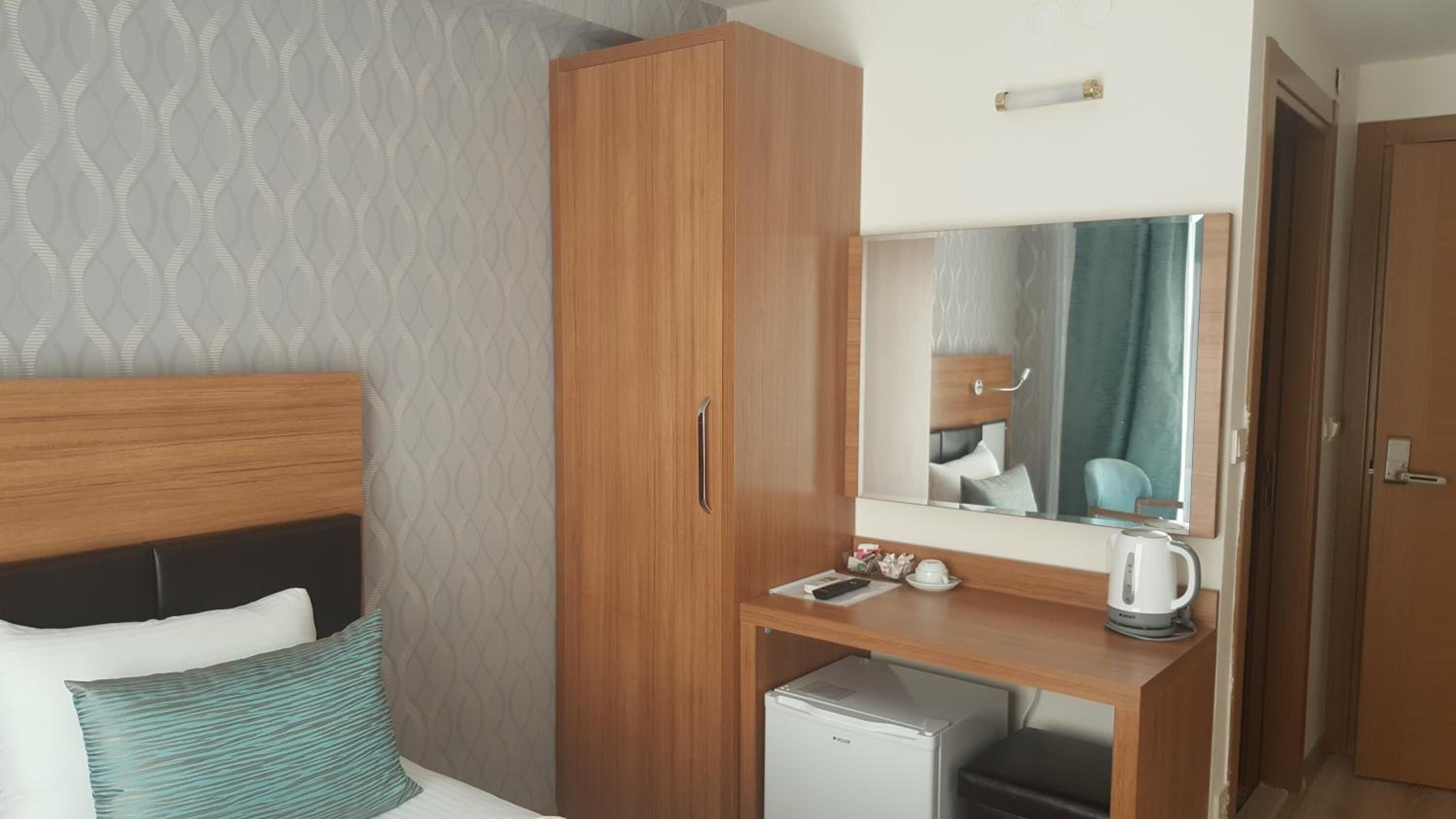 Een hotelkamer met een bed, een houten garderobe, een klein bureau met een waterkoker en een spiegel die de kamer weerspiegelt.