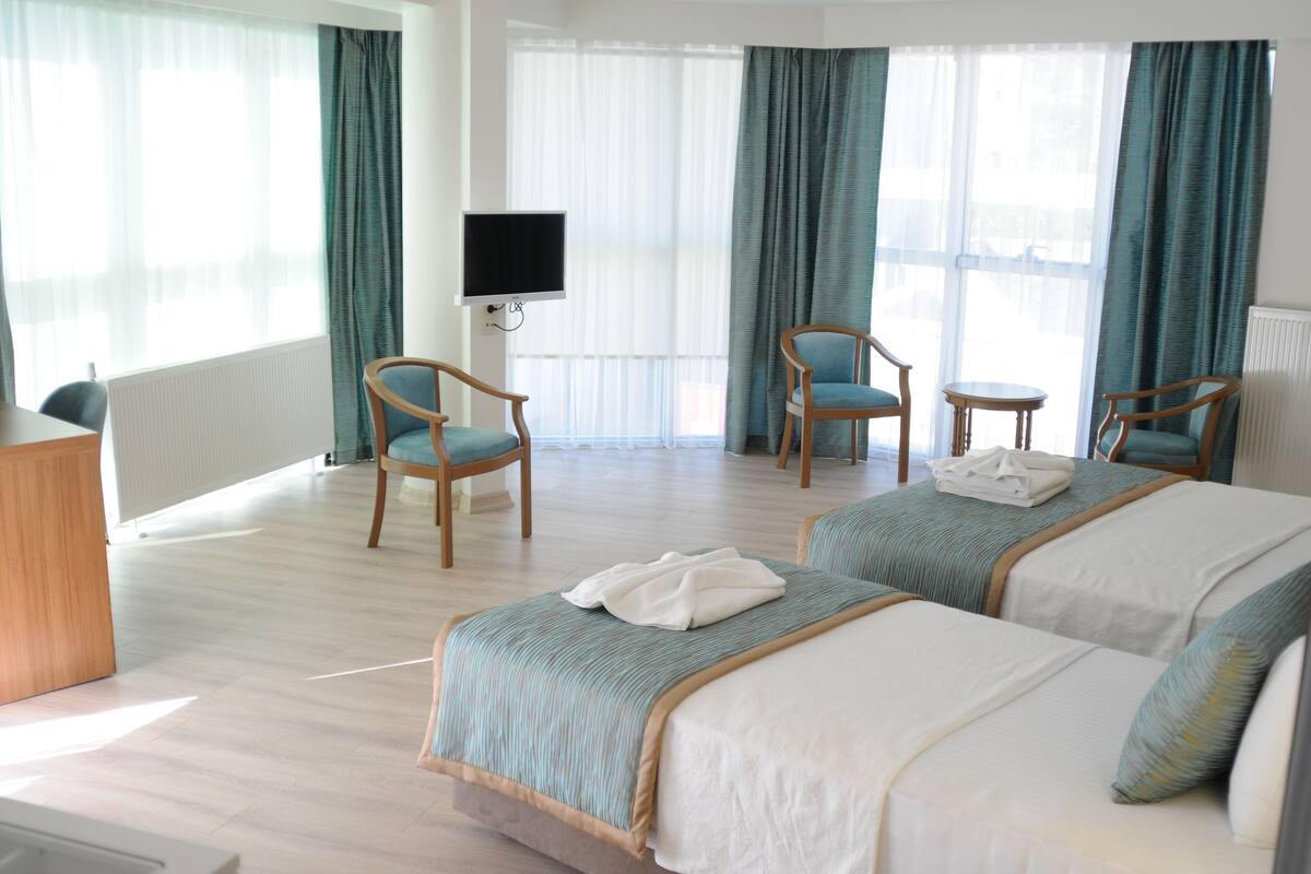 Une chambre d’hôtel avec deux lits, un bureau, une chaise, une télévision et de grandes fenêtres avec des rideaux.