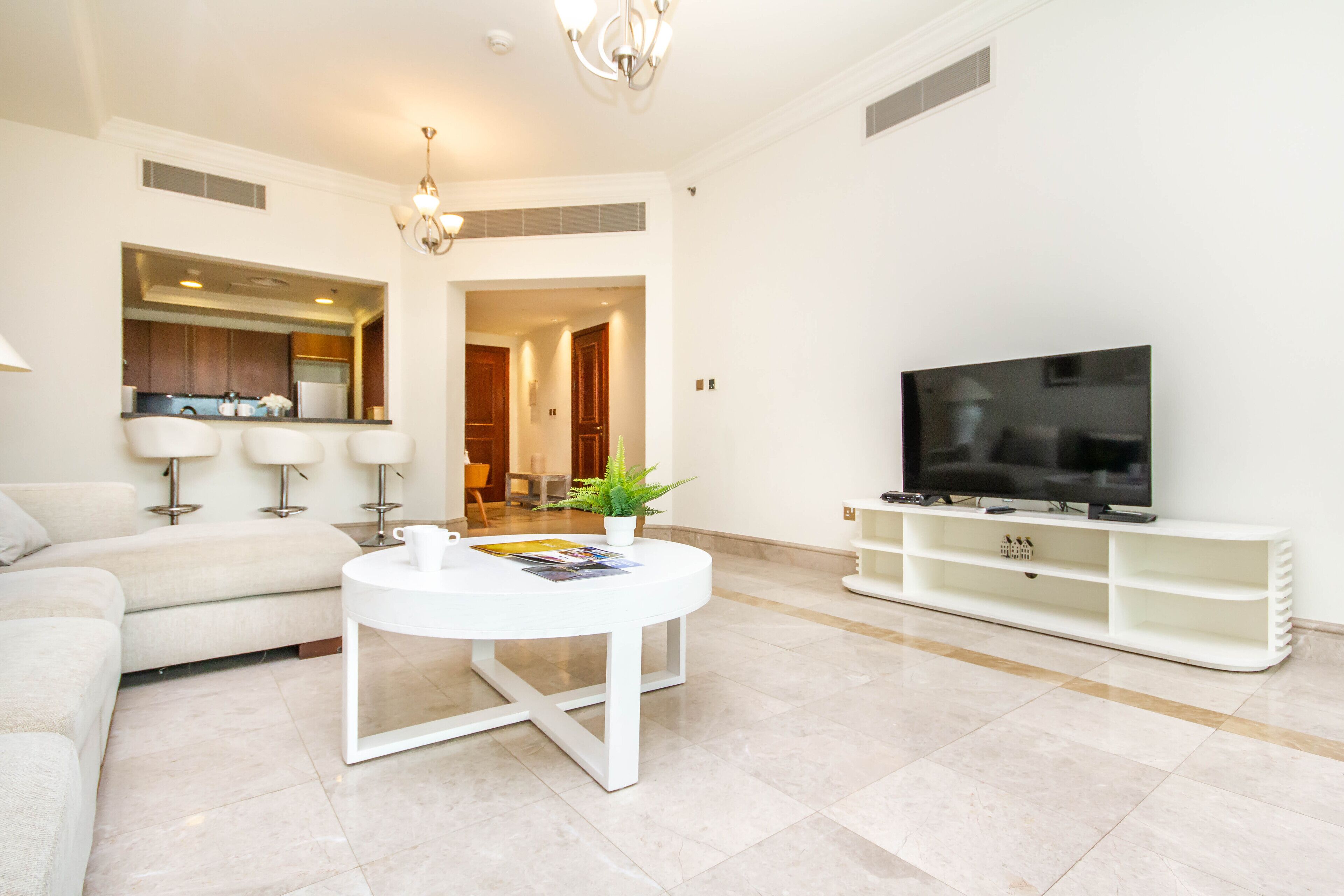 Un salon moderne avec un canapé blanc, une table basse ronde, un téléviseur à écran plat posé sur un meuble blanc, et un coin cuisine à l’arrière-plan.