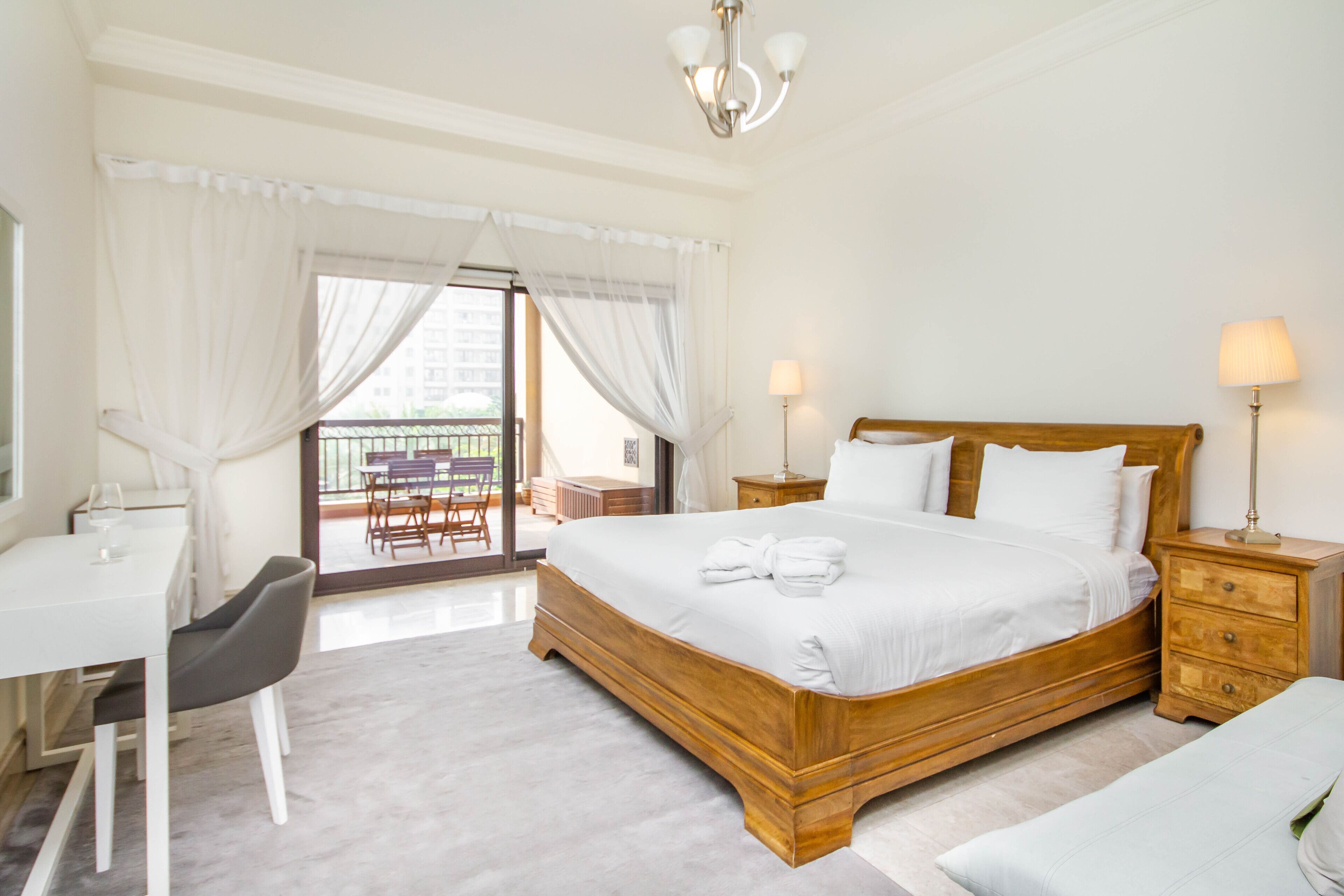 Een hotelkamer met een houten bed, witte lakens, een bureau met stoel, en een balkon met tafel en stoelen.