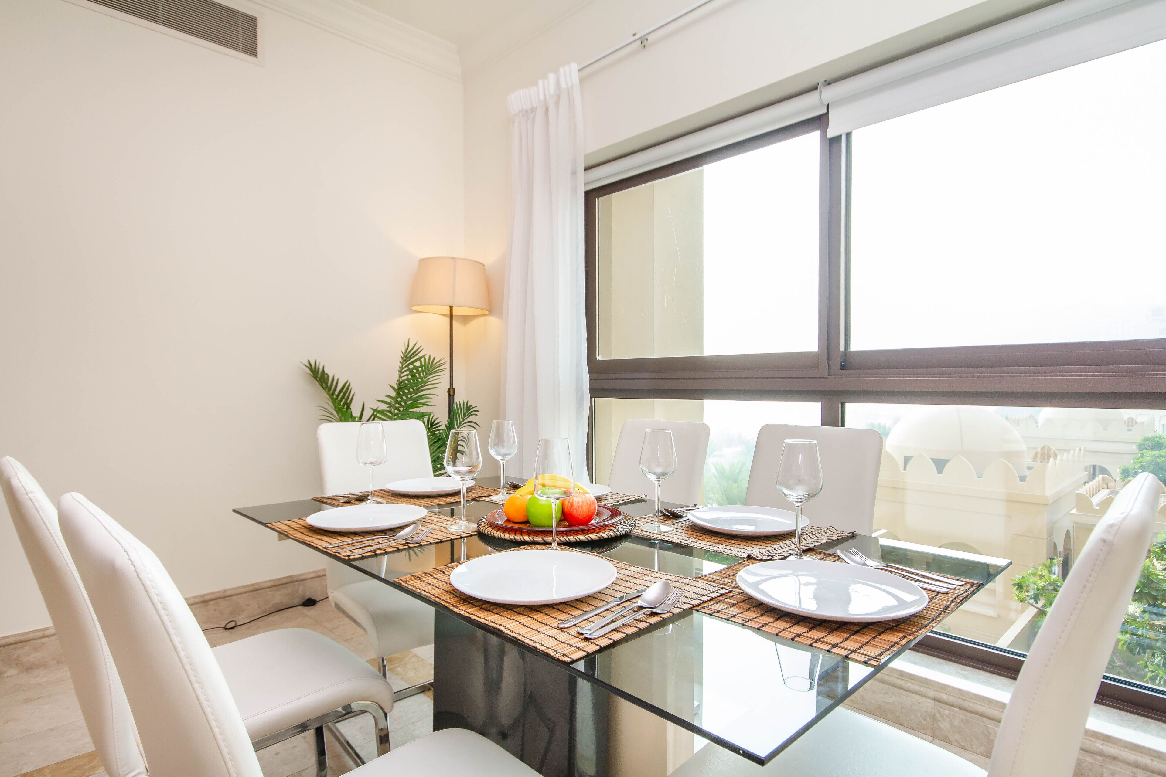Zona pranzo con tavolo in vetro, sedie bianche e vista su un edificio tramite ampie finestre.