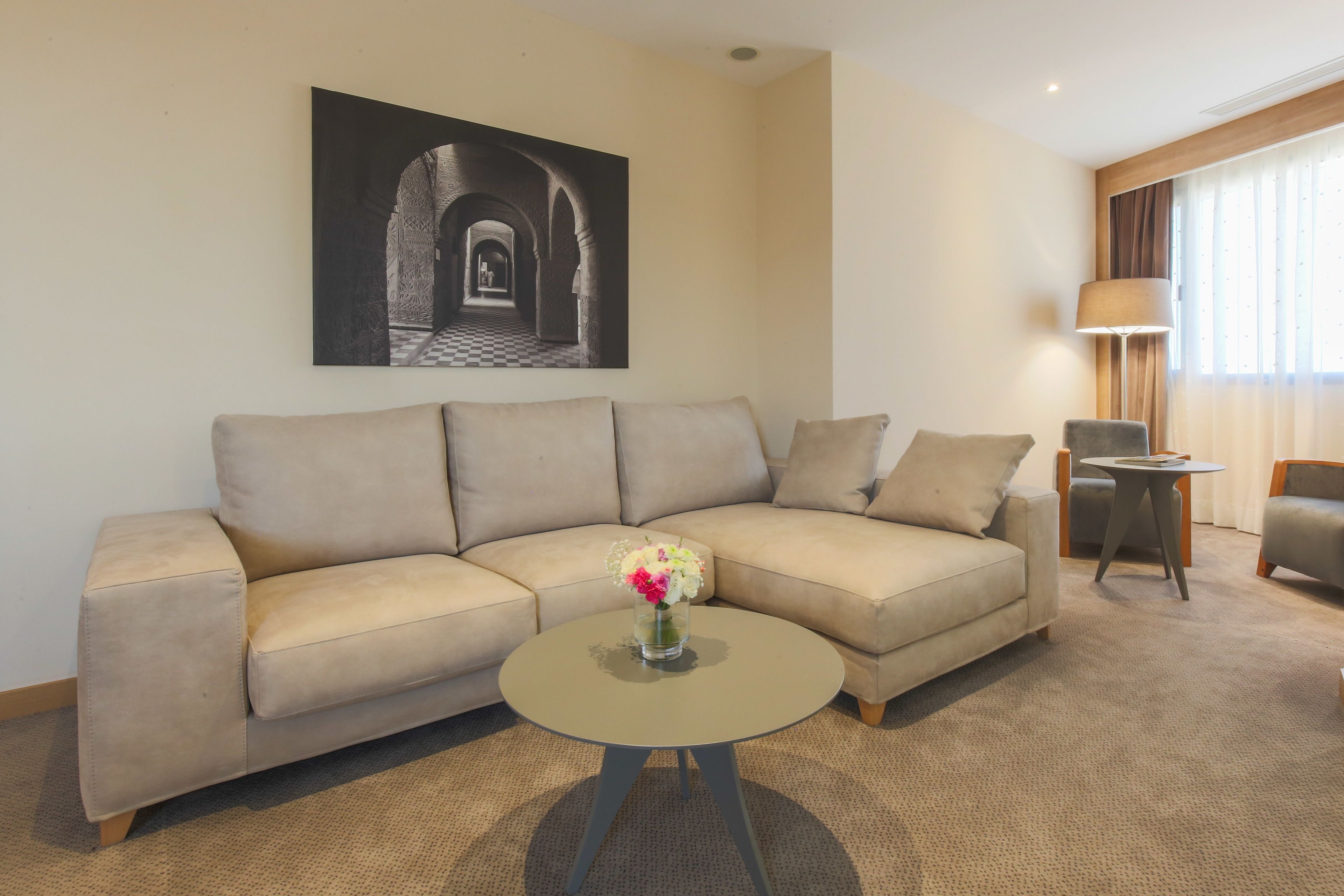 Ein modernes Wohnzimmer mit einem beigefarbenen Sofa, einem runden Couchtisch mit Blumenvase, einer Lampe und einem gerahmten Bild an der Wand.