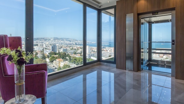 A modern hotel room with large windows offering a city view, a marble floor, and a purple chair with a vase of flowers. - Lamaraz Arts Hotel (Algiers)