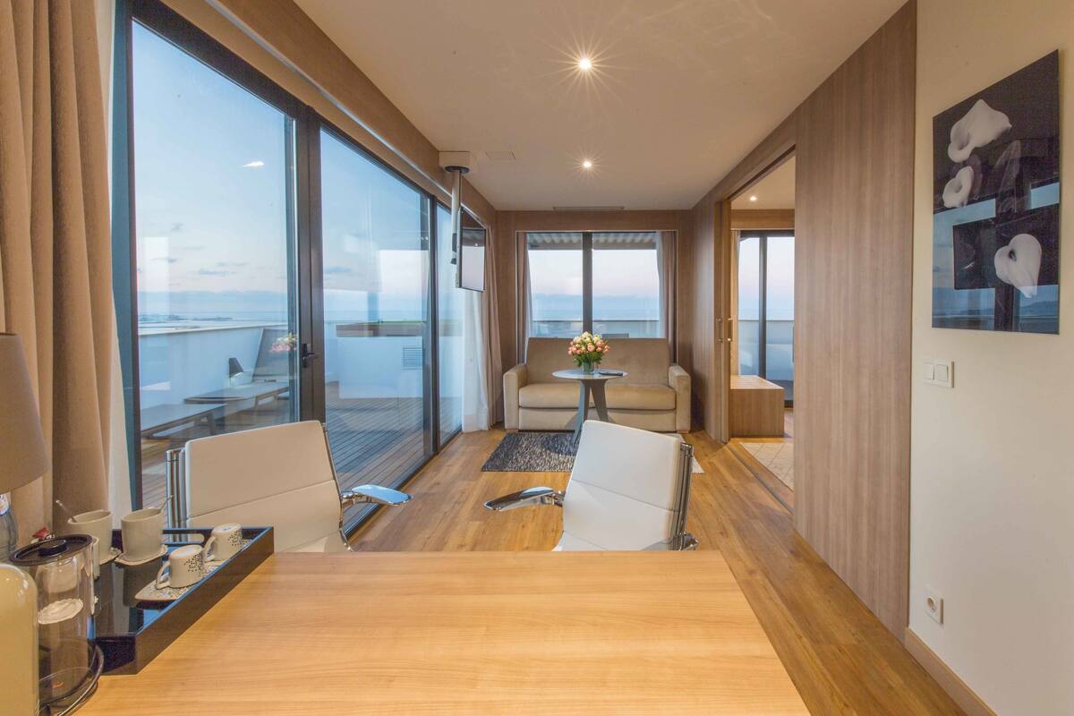 A modern hotel room with a large balcony, a seating area with a table and chairs, and a view of the ocean.