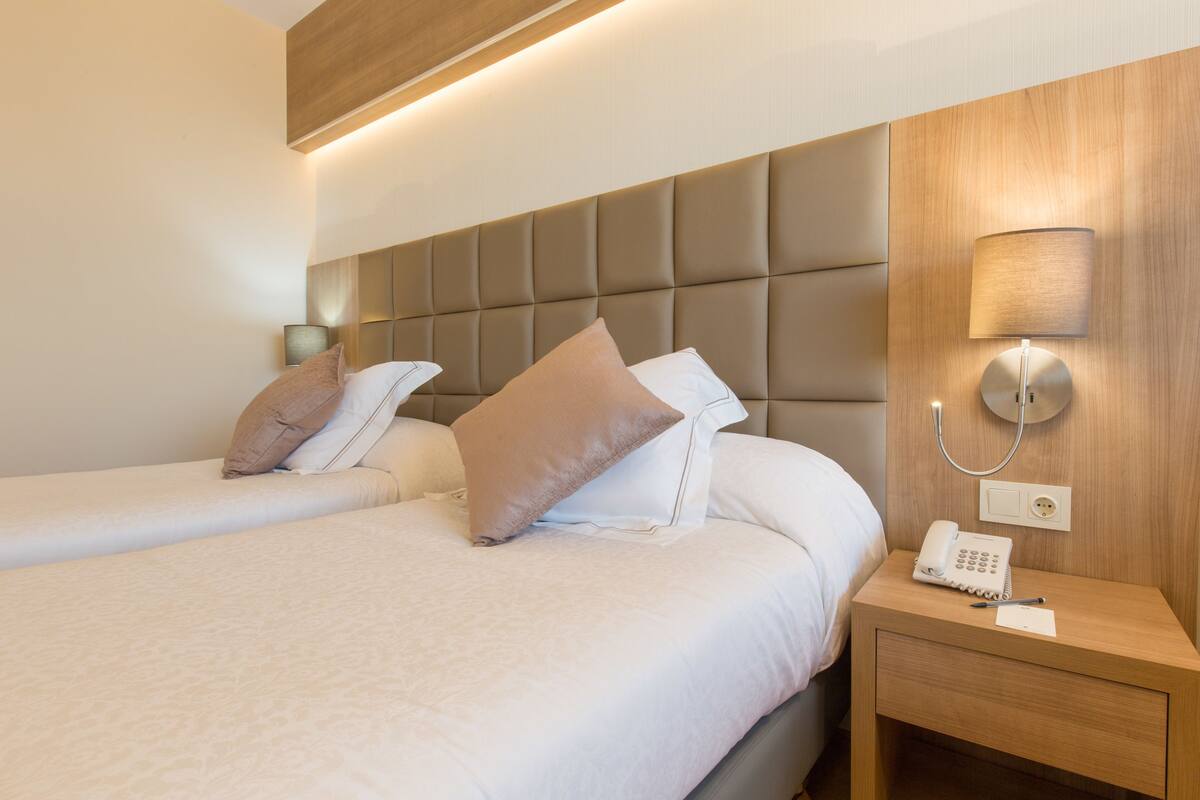 A hotel room with two beds, a bedside table with a lamp, and a phone.