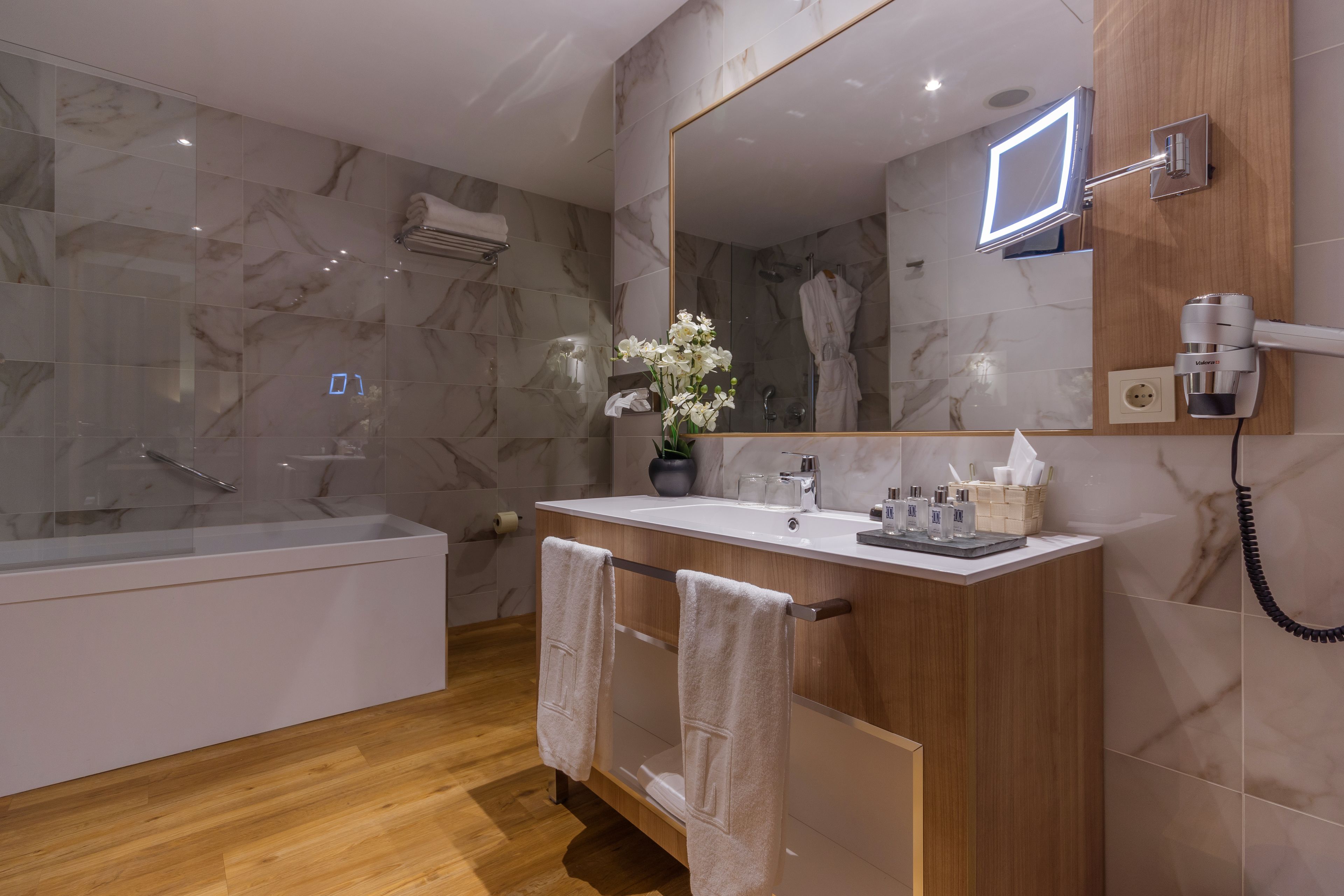 A modern bathroom with a marble wall, a wooden vanity, a large mirror, a built-in shower, and a towel rack.