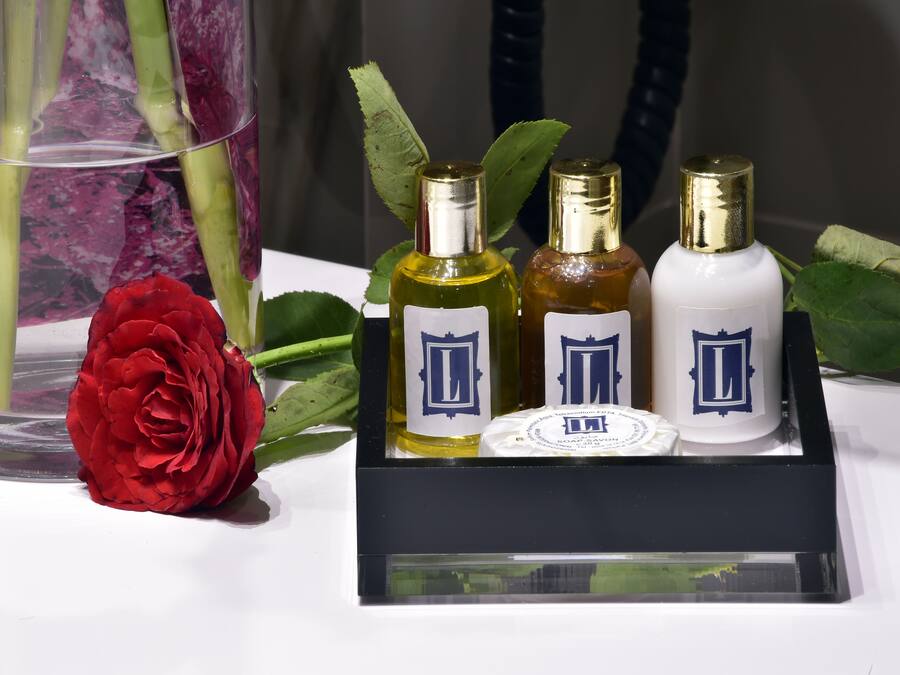 Exhibición de cuatro botellas de perfume con un recipiente jabonero redondo central, una rosa roja y hojas verdes sobre una superficie blanca.