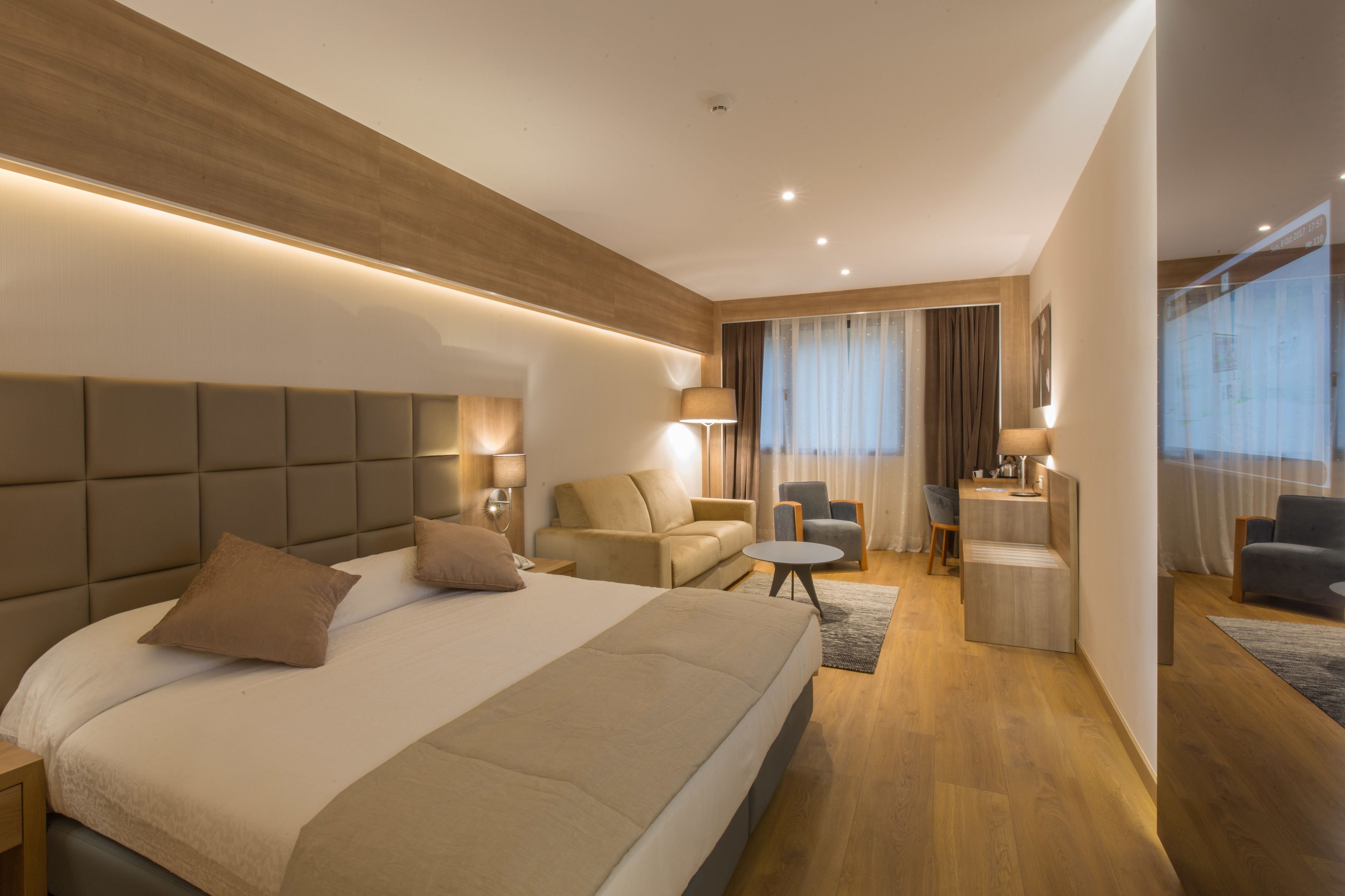 A modern hotel room with a large bed, a desk, a sofa, and a chair.