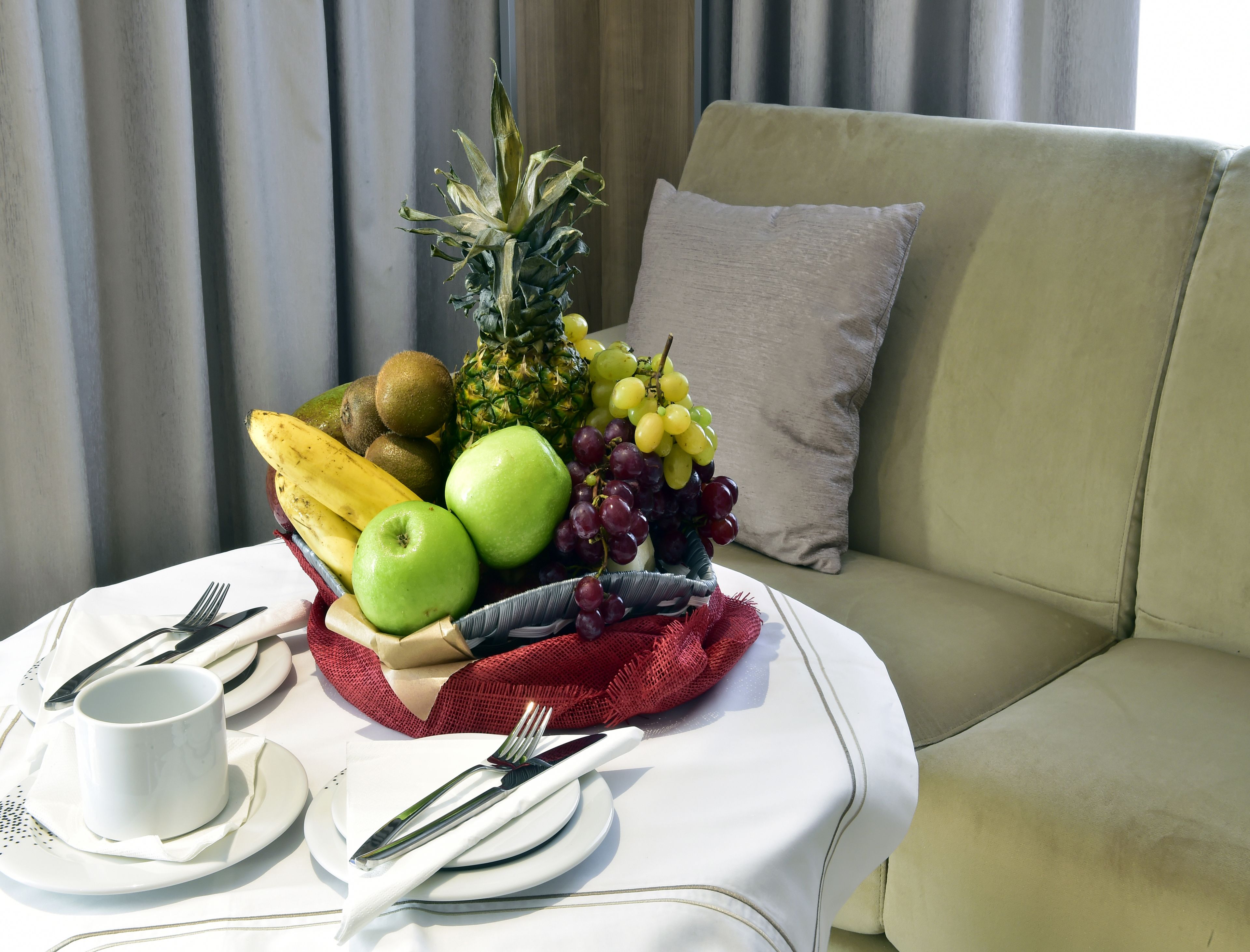 A fruit basket on a table with a white tablecloth, a cup, and cutlery.