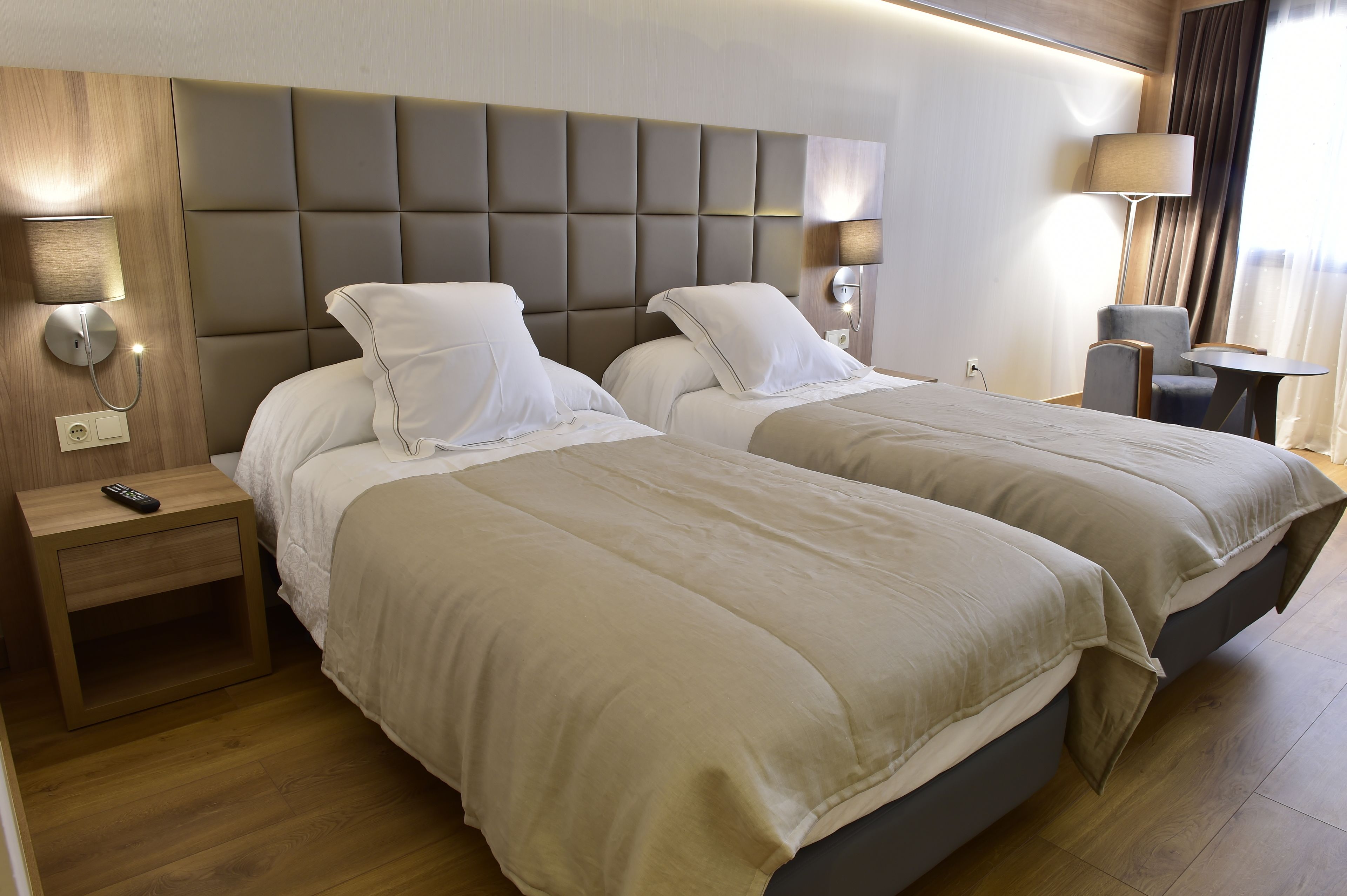 A hotel room with two beds, a nightstand with a remote control, a lamp, and a small table.