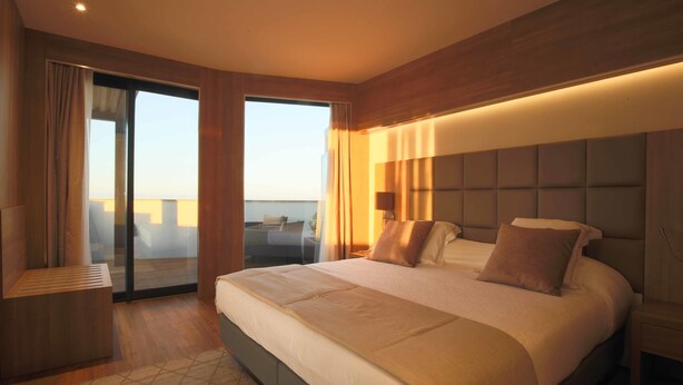 A modern hotel room with a large bed, a seating area with a view, and a sliding glass door to the outside.