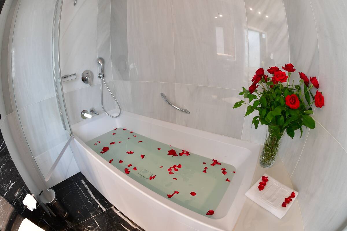 A modern bathroom with a glass-enclosed shower, a bathtub with rose petals, and a vase of red roses.