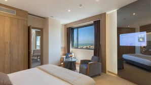 A hotel room with a large bed, a desk, two chairs, a small table with a vase, and a large window with a view of the sea. - Lamaraz Arts Hotel (Algiers)