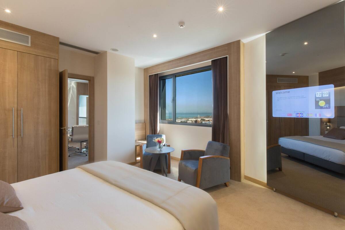 A hotel room with a large bed, a desk, two chairs, a small table with a vase, and a large window with a view of the sea.