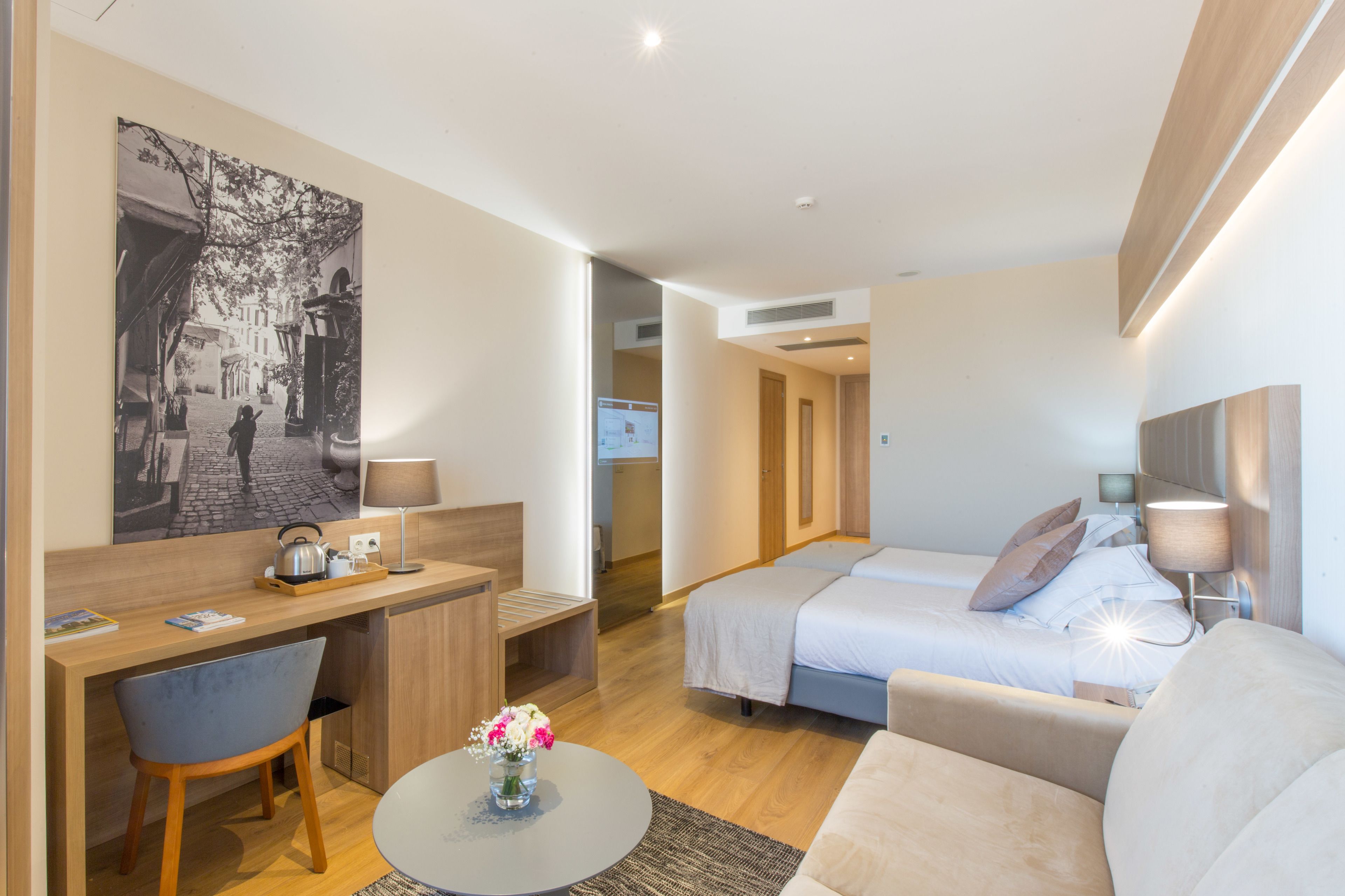 A modern hotel room with a bed, a desk, a chair, and a small table with a vase of flowers.