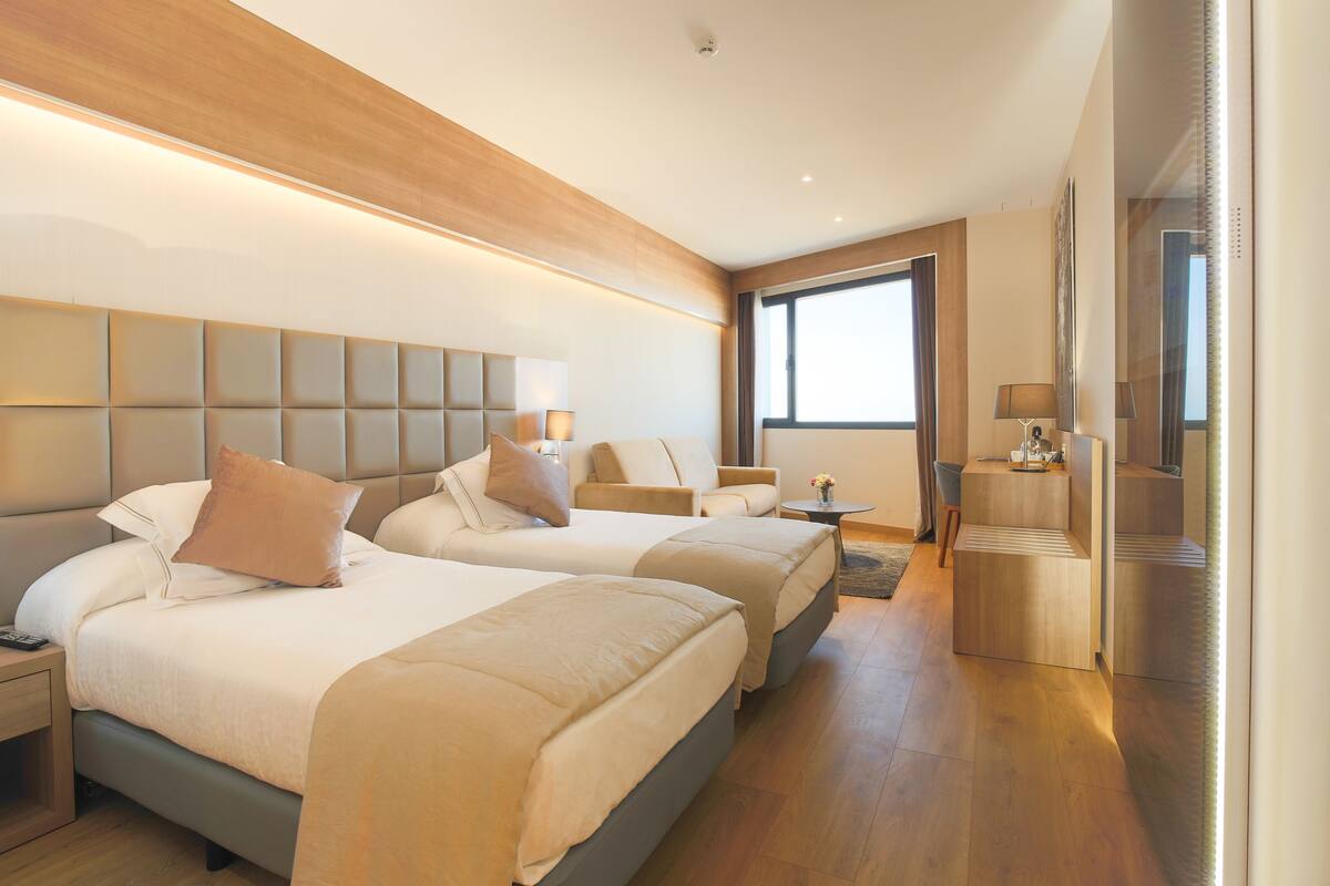 A modern hotel room with two beds, a sitting area, and a large window.