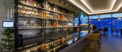 A modern bar with a high bar counter, shelves of liquor bottles, and a city view through large windows.