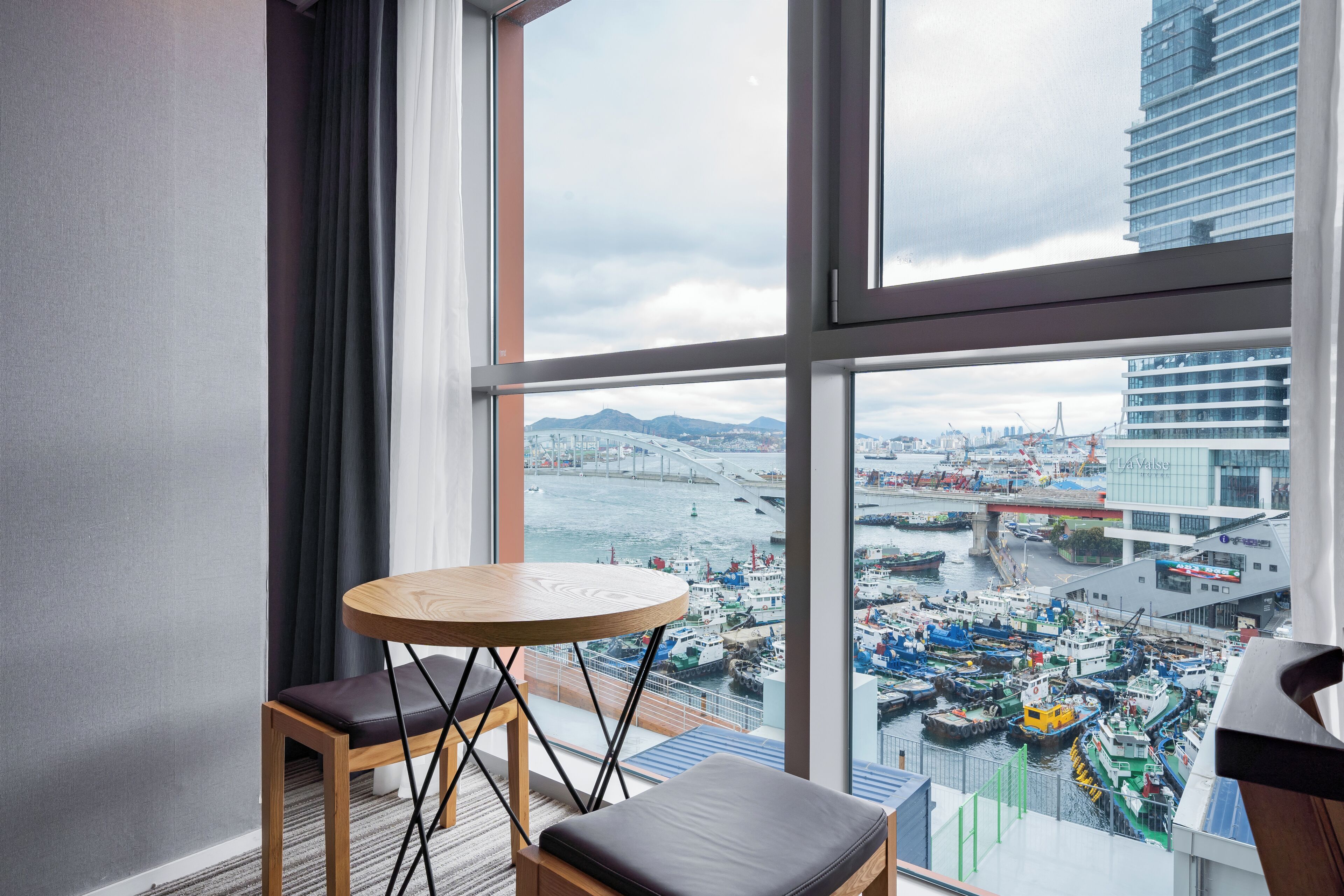 A modern hotel room with a large window offering a view of a busy port and cityscape.