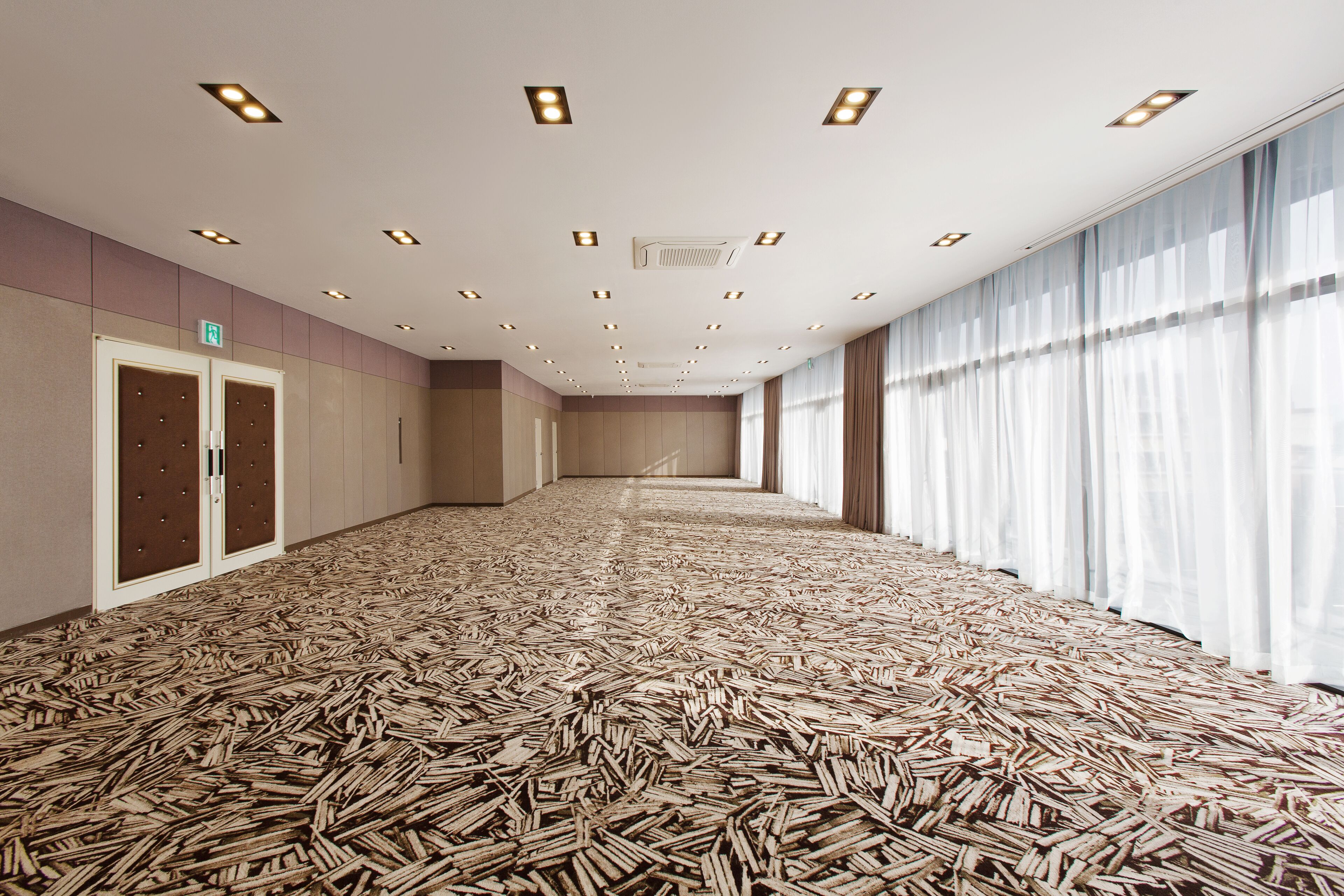 A spacious hotel corridor with a patterned carpet, recessed lighting, and large windows with sheer curtains.