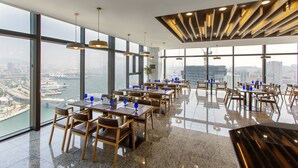En restaurant i en høj bygning med store vinduer, der giver udsigt over byen, skinnende gulve og moderne møbler i træ.