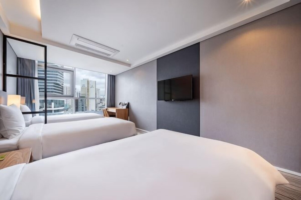 A hotel room with two beds, a desk, a chair, a TV, and a large window with a city view.