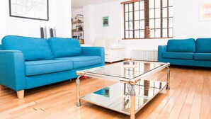 Uma sala de estar com dois sofás azuis, uma mesa de centro de vidro, piso de madeira, um radiador e uma janela com persianas.