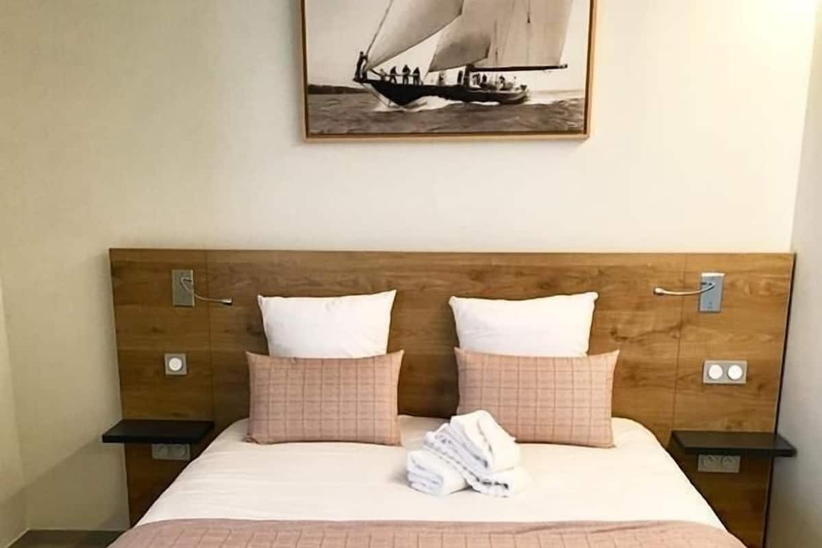 Una cama bien tendida con un edredón rosa, dos mesitas de noche de madera con lámparas, y un cuadro en la pared con una imagen de un velero.