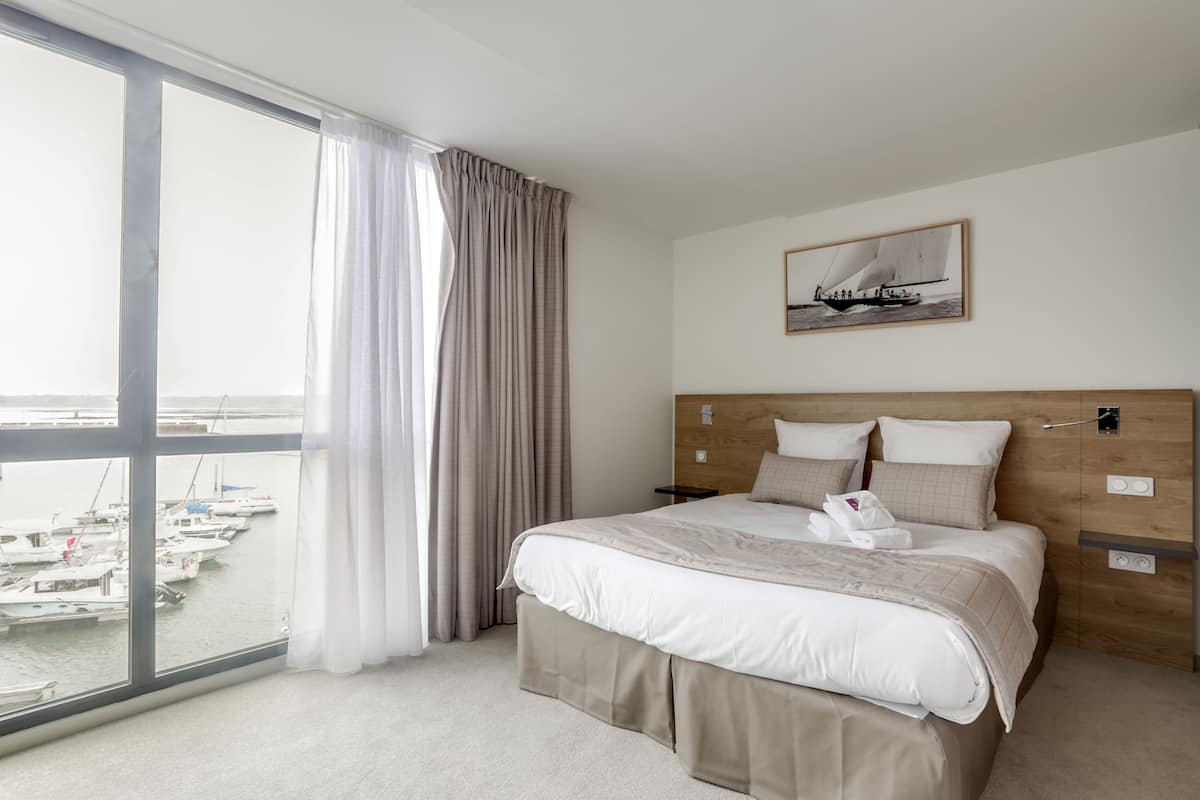 Habitación de hotel con un ventanal grande, una cama con cabecero de madera y un cuadro enmarcado en la pared.