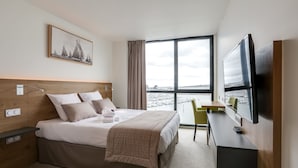 A modern hotel room with a bed, a desk, a television, and a large window offering a view of a marina. - Le Nautica by Akena Hotels (Perros-Guirec)