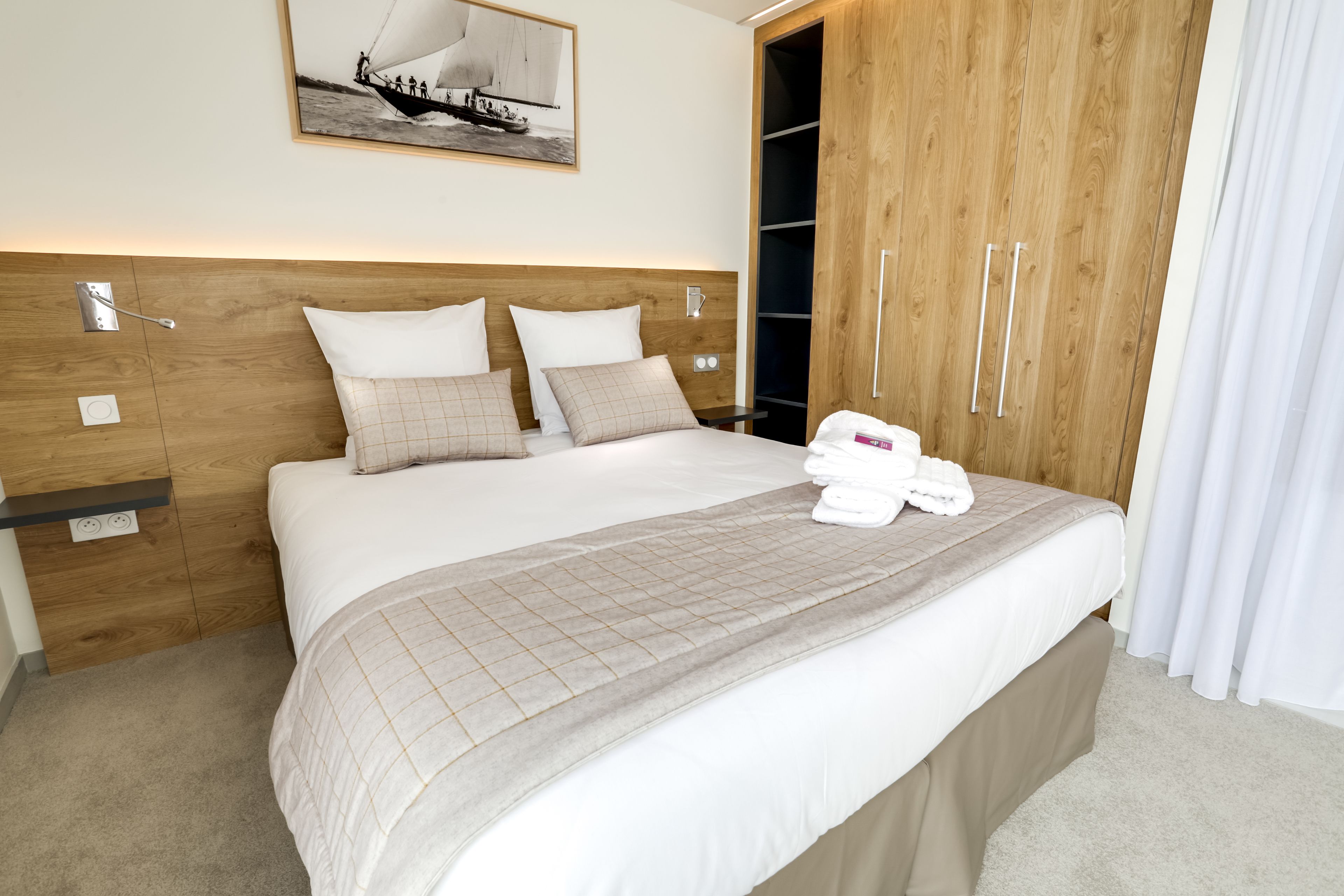 Una cama bien hecha con colcha, almohadas y una toalla doblada. Se observa un armario de madera con espejo y repisa.