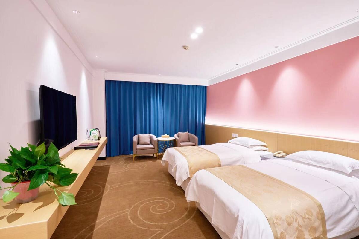 A hotel room with two beds, a desk with a chair, a TV, and a large window with blue curtains.