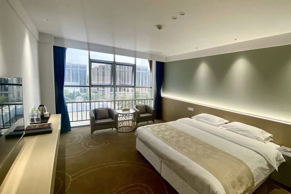 A hotel room with a large bed, a desk with a chair, a small round table, and a view of the city through large windows.