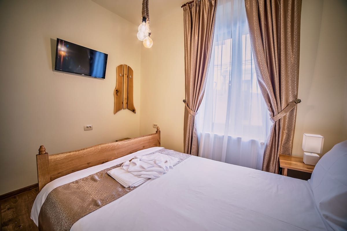 A neatly made bed with white linens, a mounted flat-screen TV, a wooden headboard, a bedside table with a lamp, and a window with curtains.