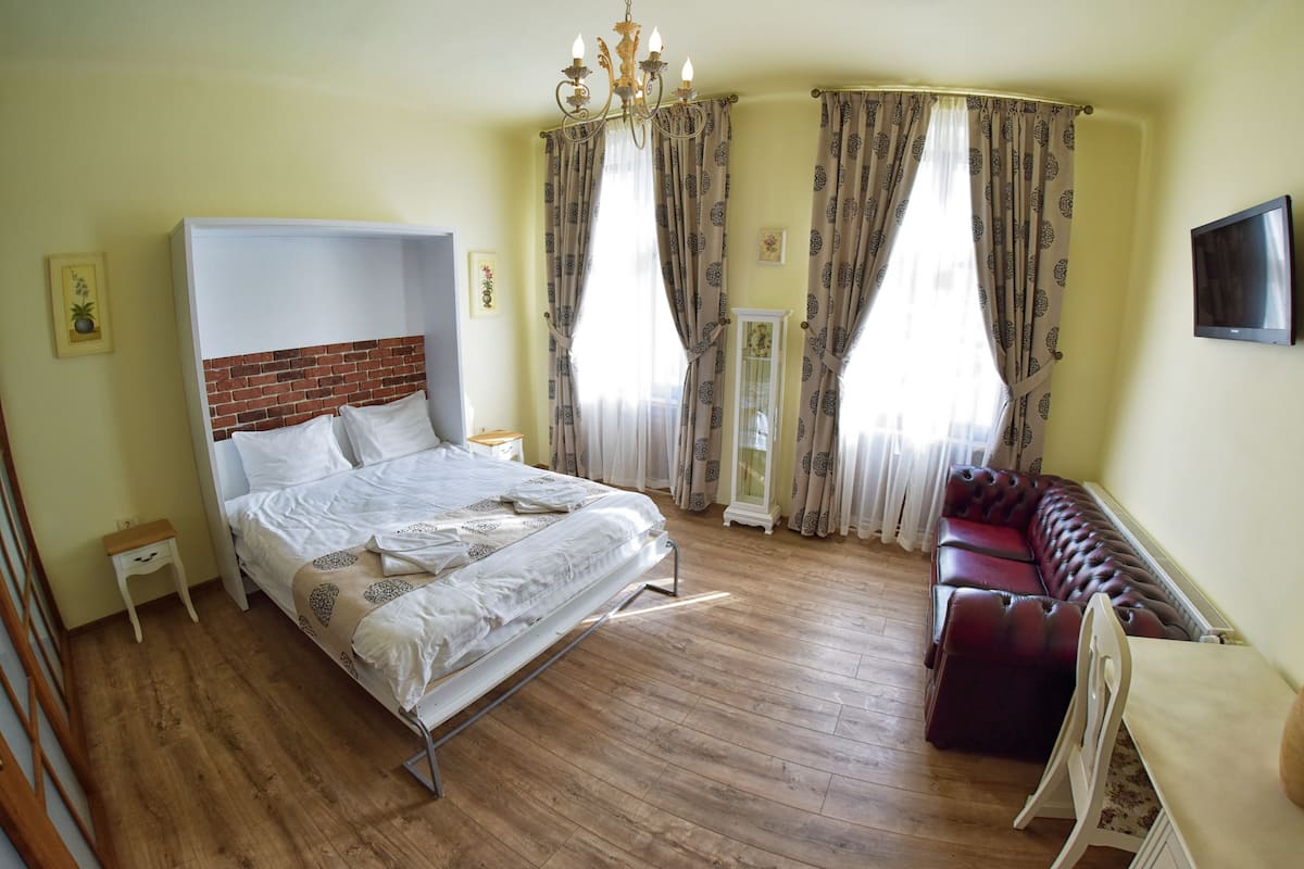 A bedroom with a bed, a small bedside table, a nightstand, a sofa, a TV, and a window with curtains.