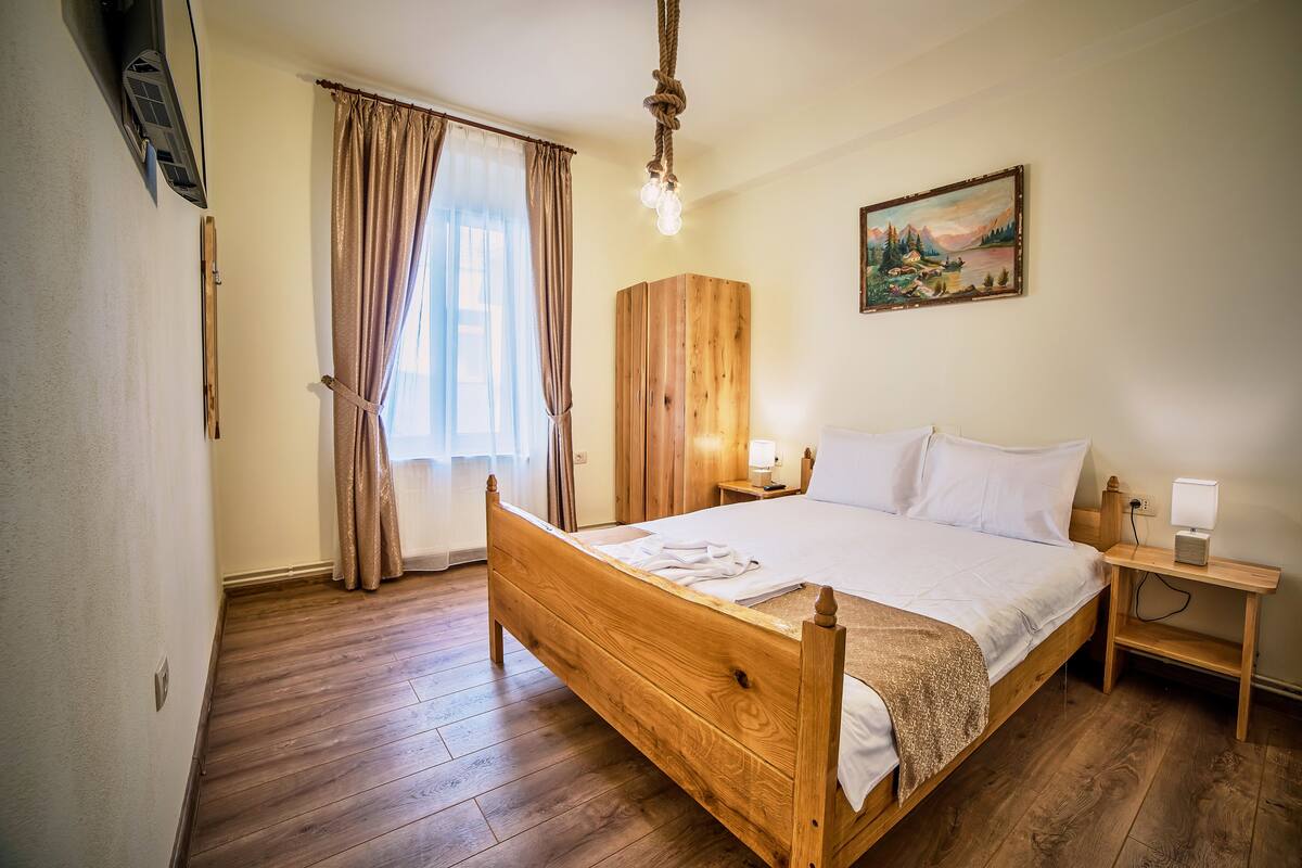 A wooden bed with white linens, a bedside table with a lamp, a painting on the wall, and a window with curtains.