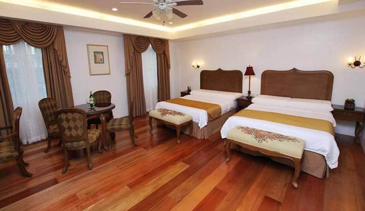 A hotel room with two beds, a dining table with chairs, a ceiling fan, and a window with curtains.