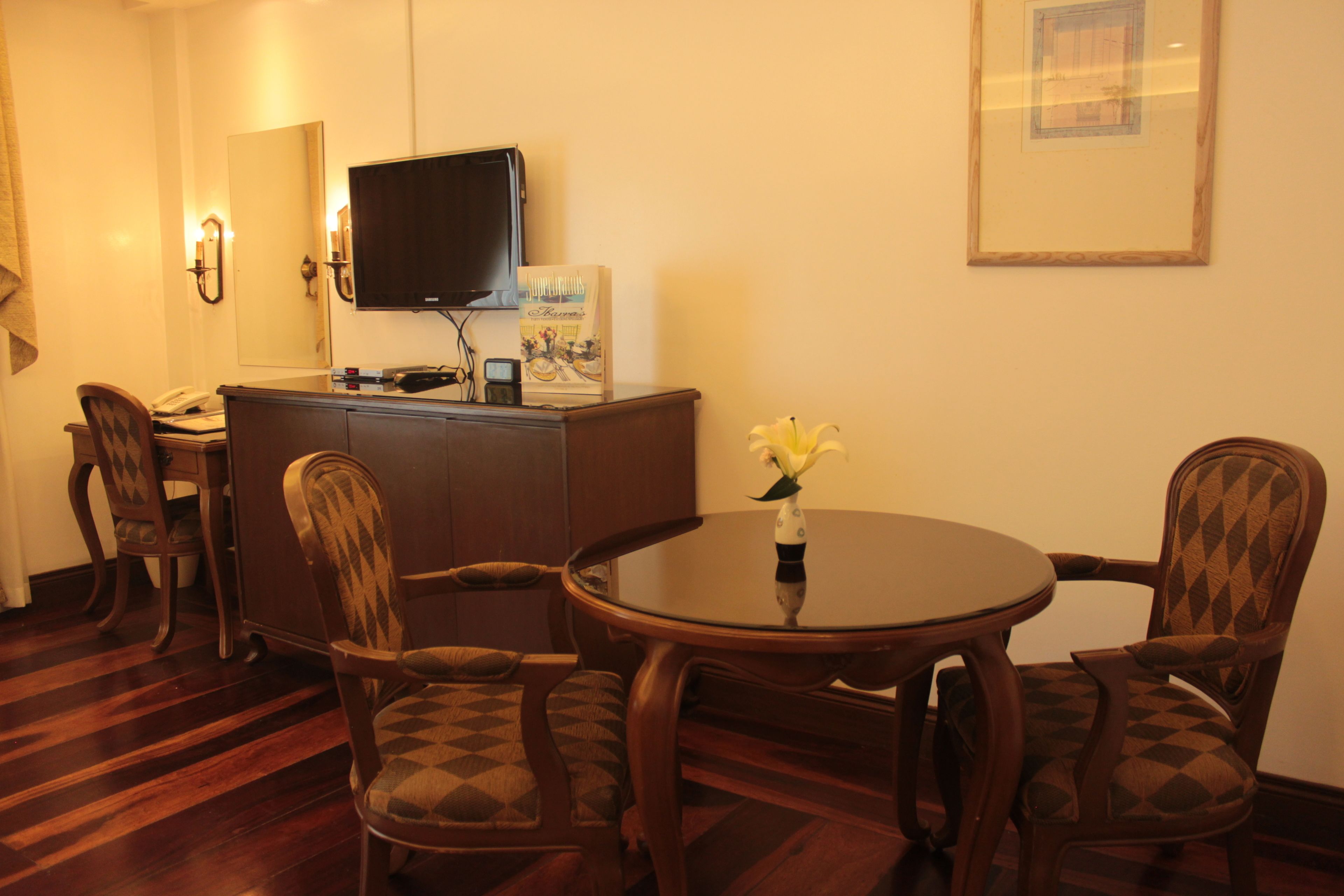 A hotel room with a wooden dining table, four patterned chairs, a small round table with a vase of flowers, a television on a cabinet, and a framed picture on the wall.