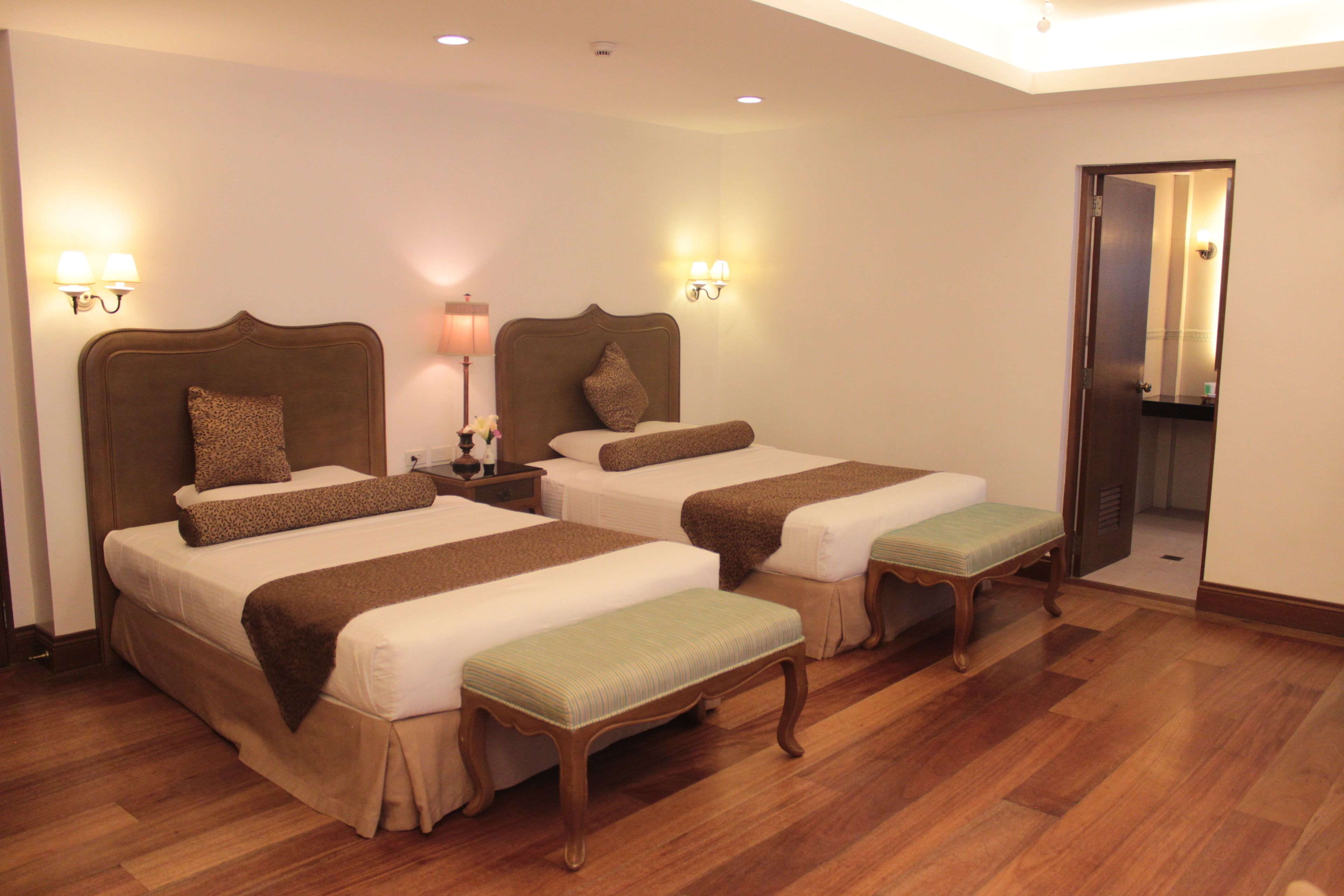 A hotel room with two beds, wooden flooring, and wall-mounted lamps.