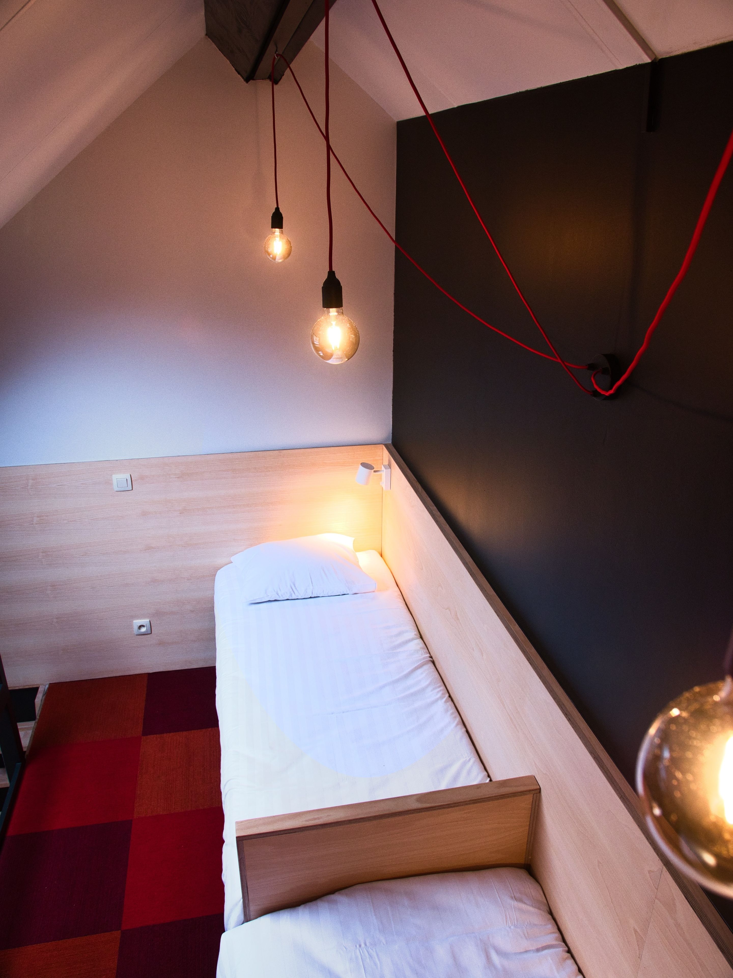 A compact, modern room with a single bed, a red and white checkered rug, and exposed wooden beams on the ceiling.